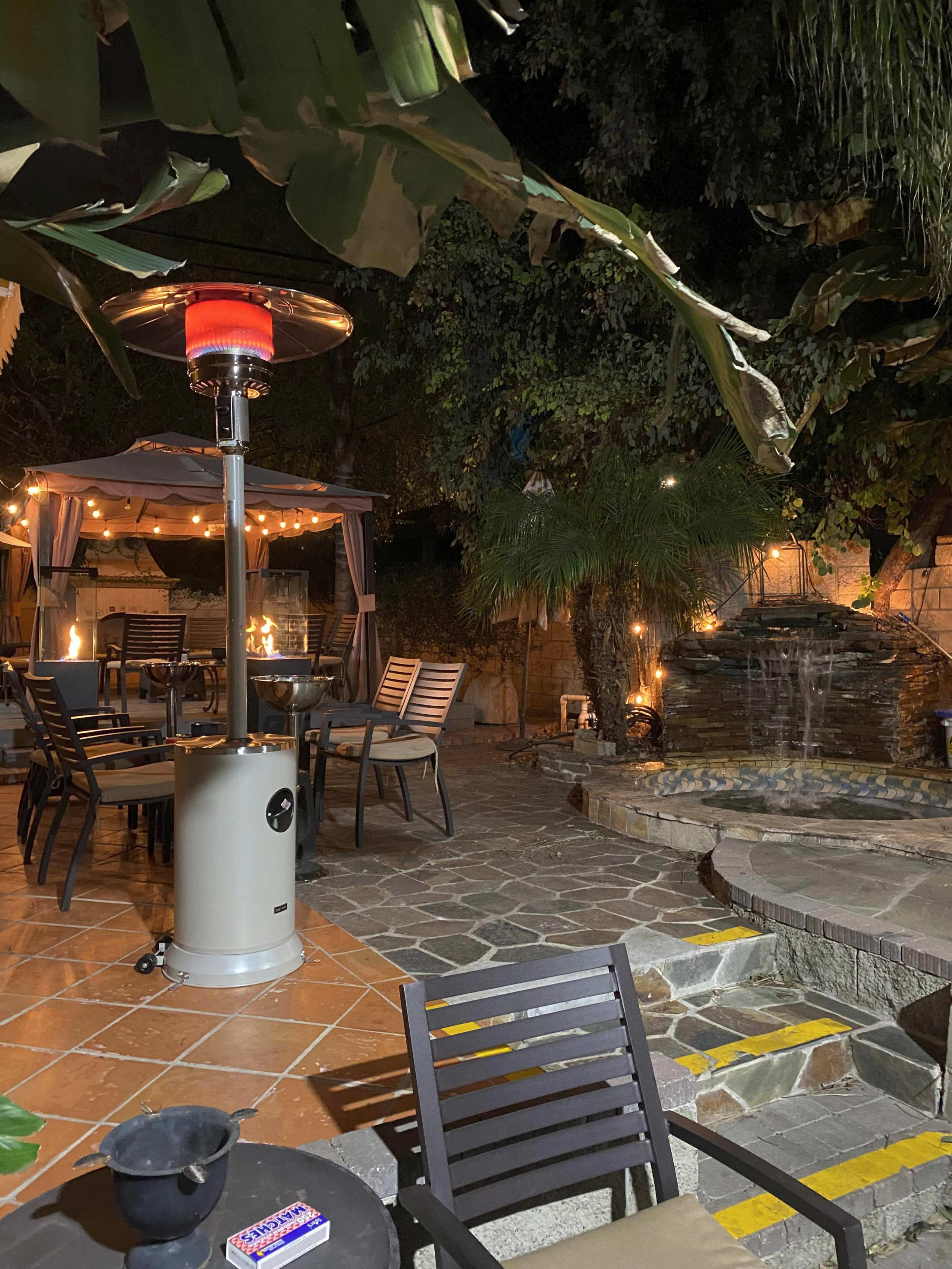 A patio area features a stone fountain, outdoor seating, a gas heater, and decorative string lights under a canopy of trees.