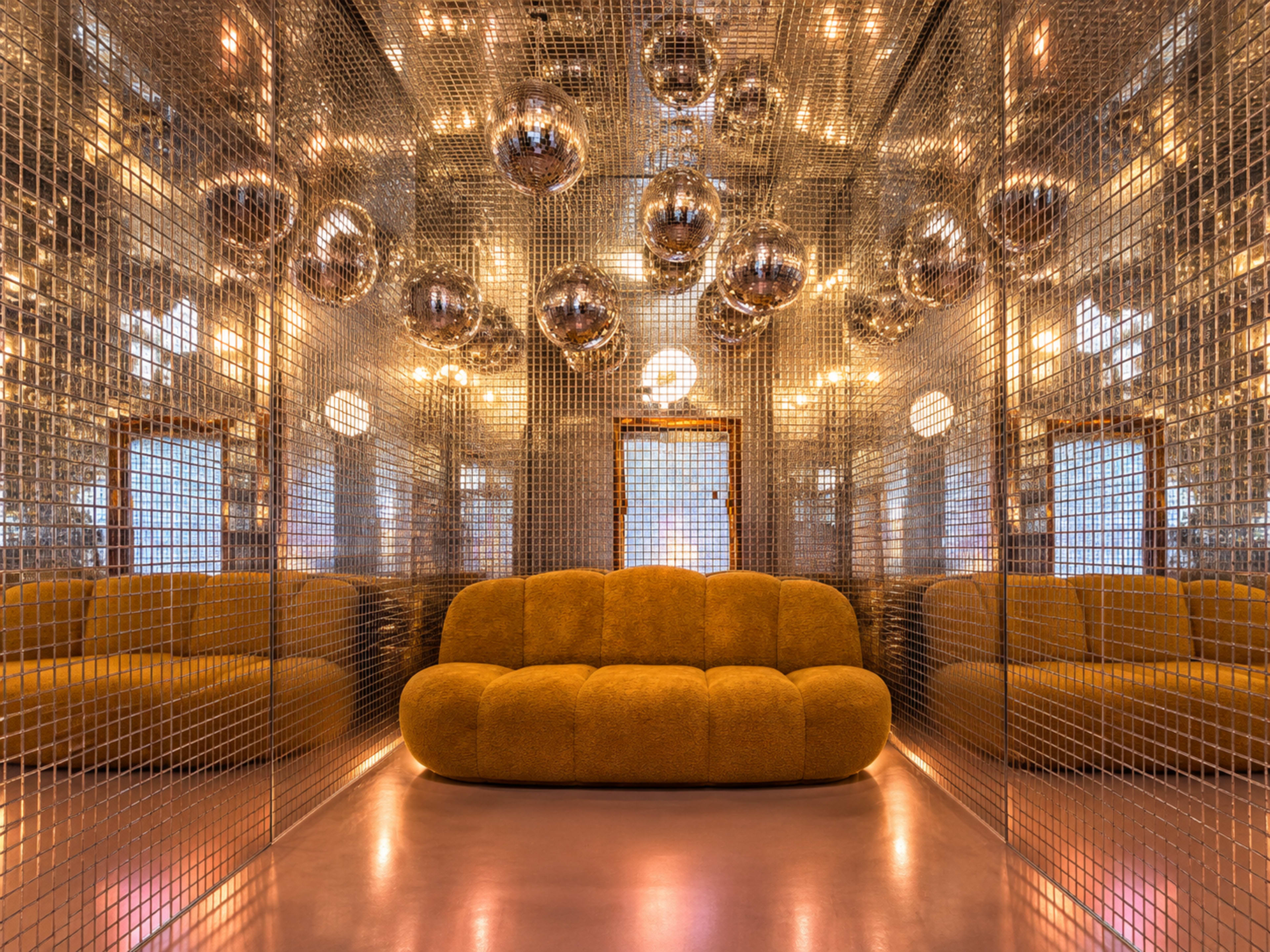 A modern lounge features a golden, curved sofa surrounded by mirrored walls and spherical light fixtures.
