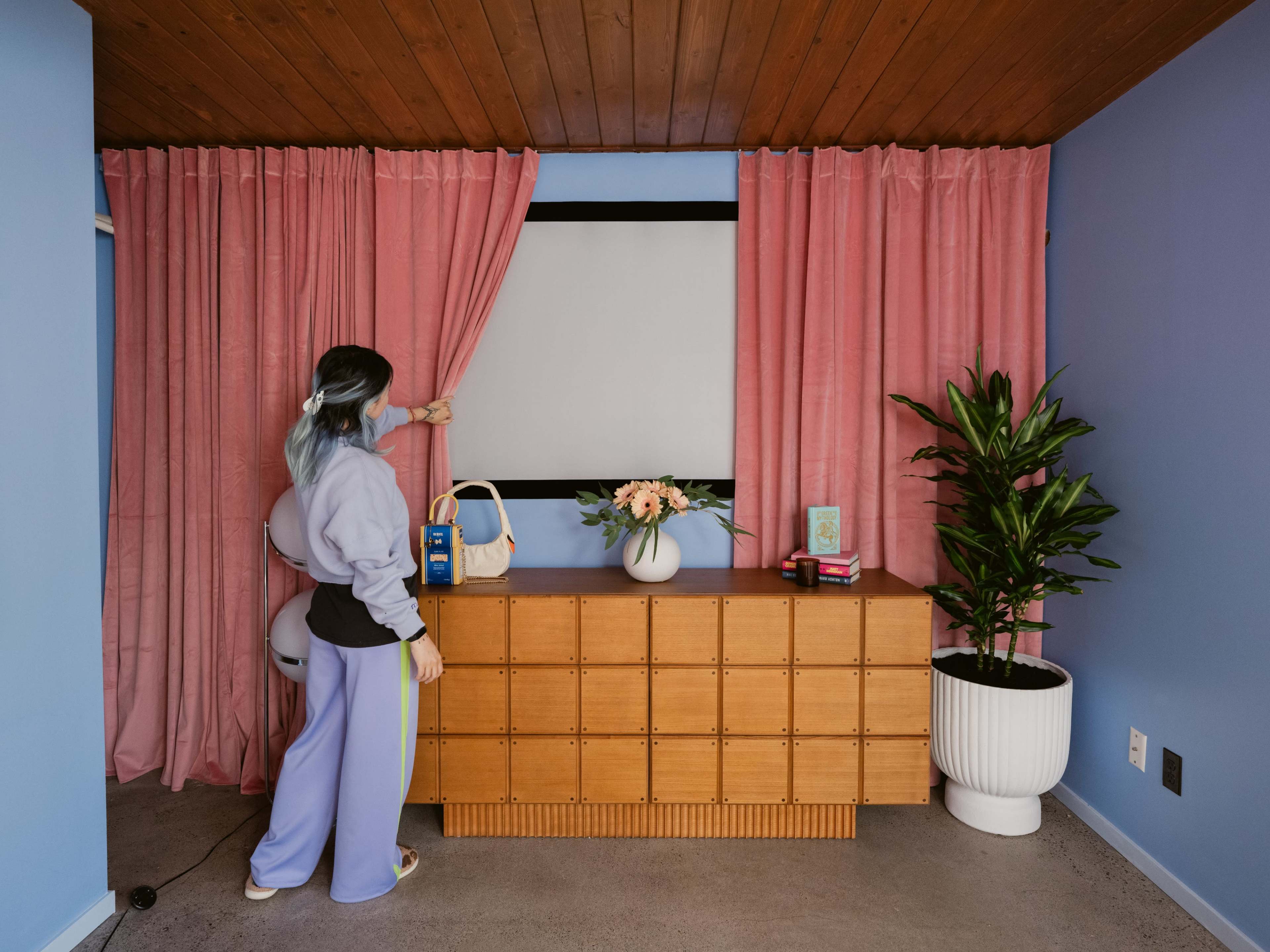 A person with blue-tinted hair is pulling back pink curtains to reveal a blank screen in a room featuring a wooden dresser and a potted plant.