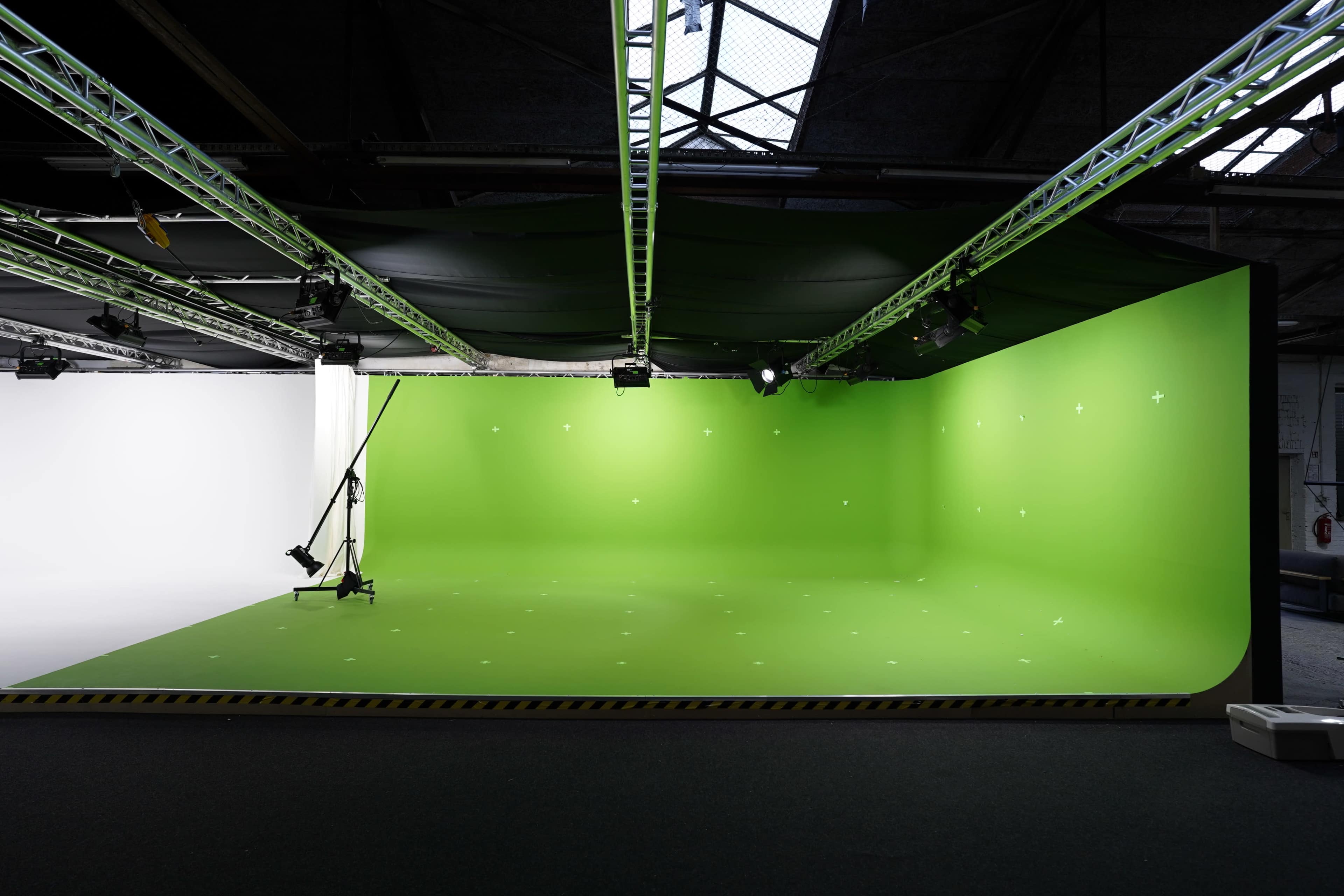 The image shows a studio space with a bright green backdrop and a white backdrop, equipped with lighting and a camera support system.