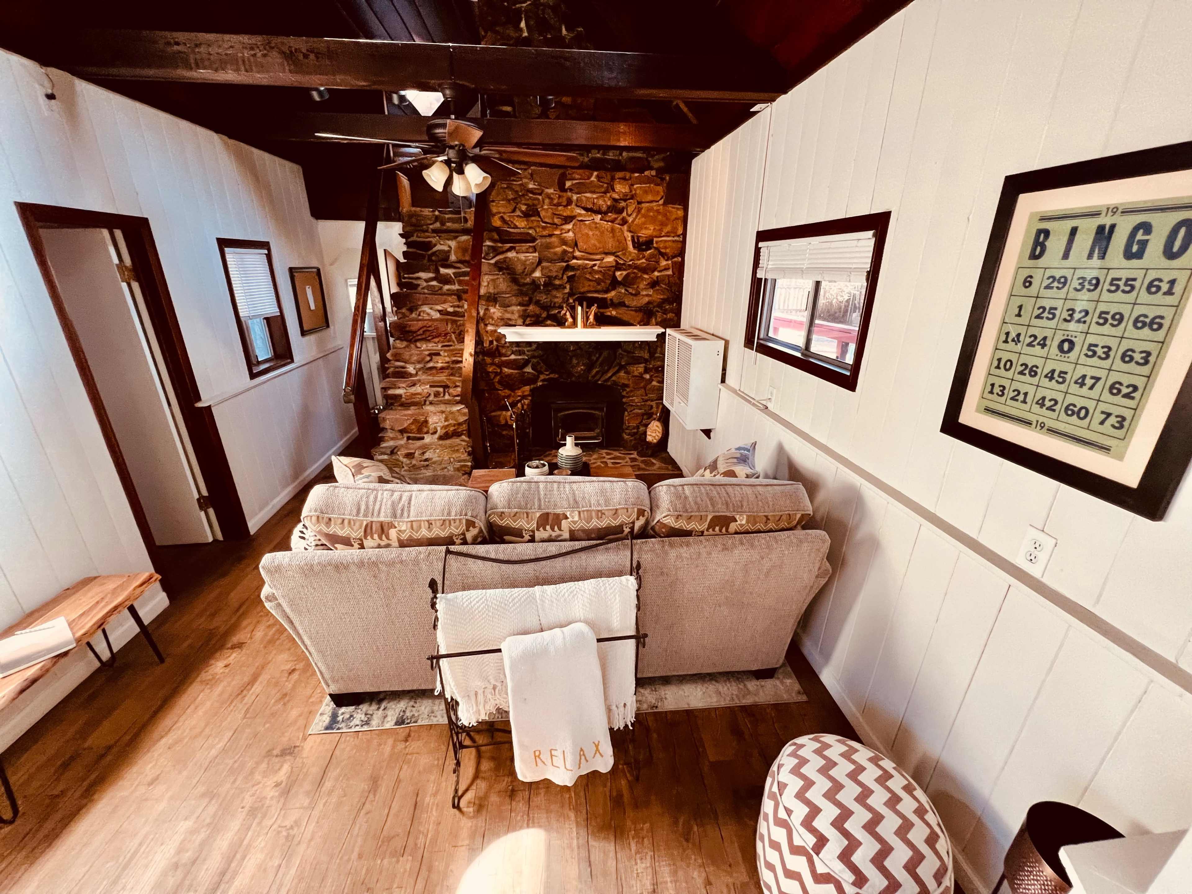 A cozy living room with a stone fireplace, wooden beams, and a seating area featuring a beige couch and a patterned ottoman.