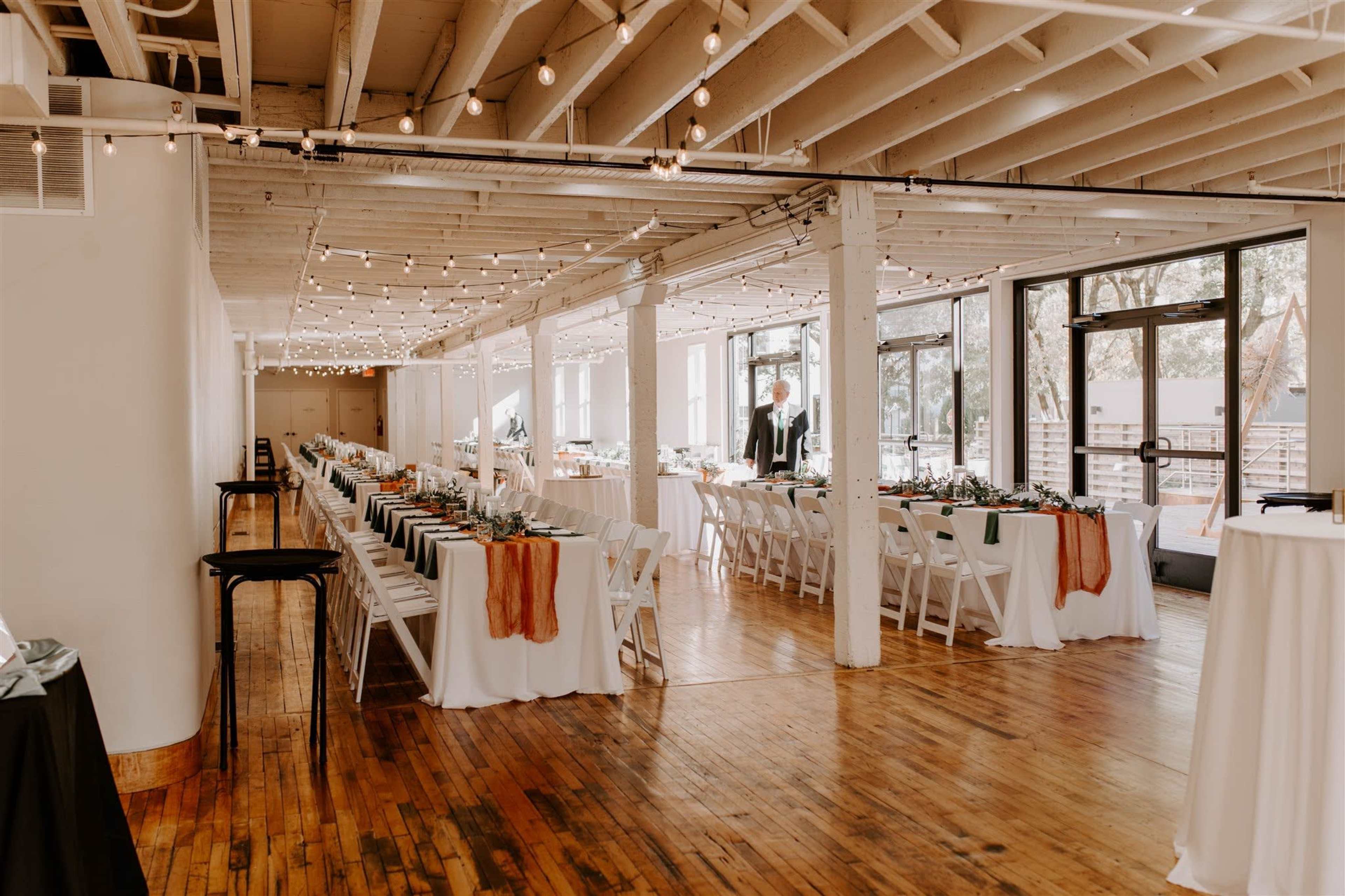 The image shows a large event space with long tables set for a meal, featuring white tablecloths and string lights overhead.