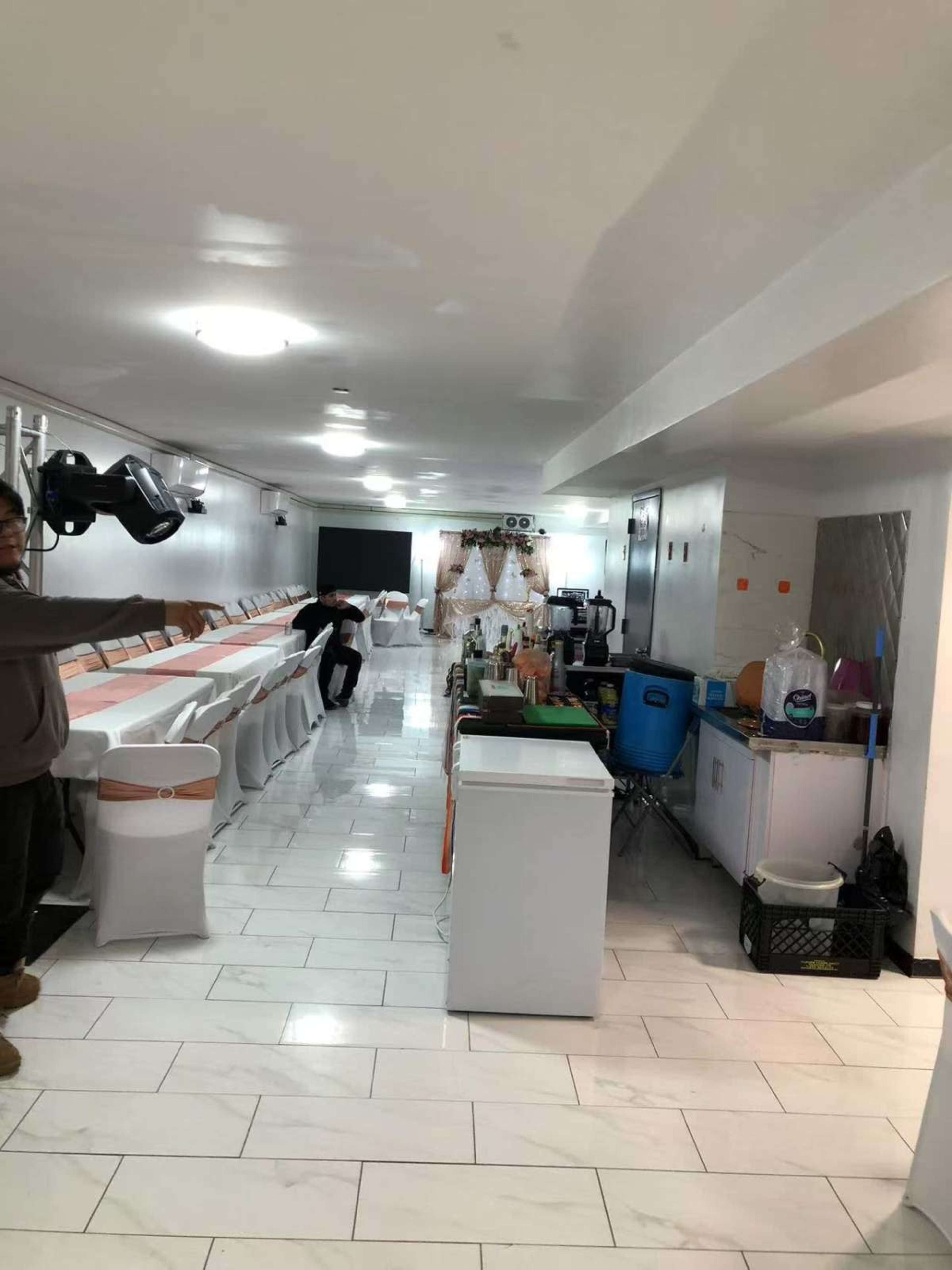 The image shows a banquet hall setup with chairs and tables arranged along the walls, a serving area containing appliances, and decorative elements at the far end.