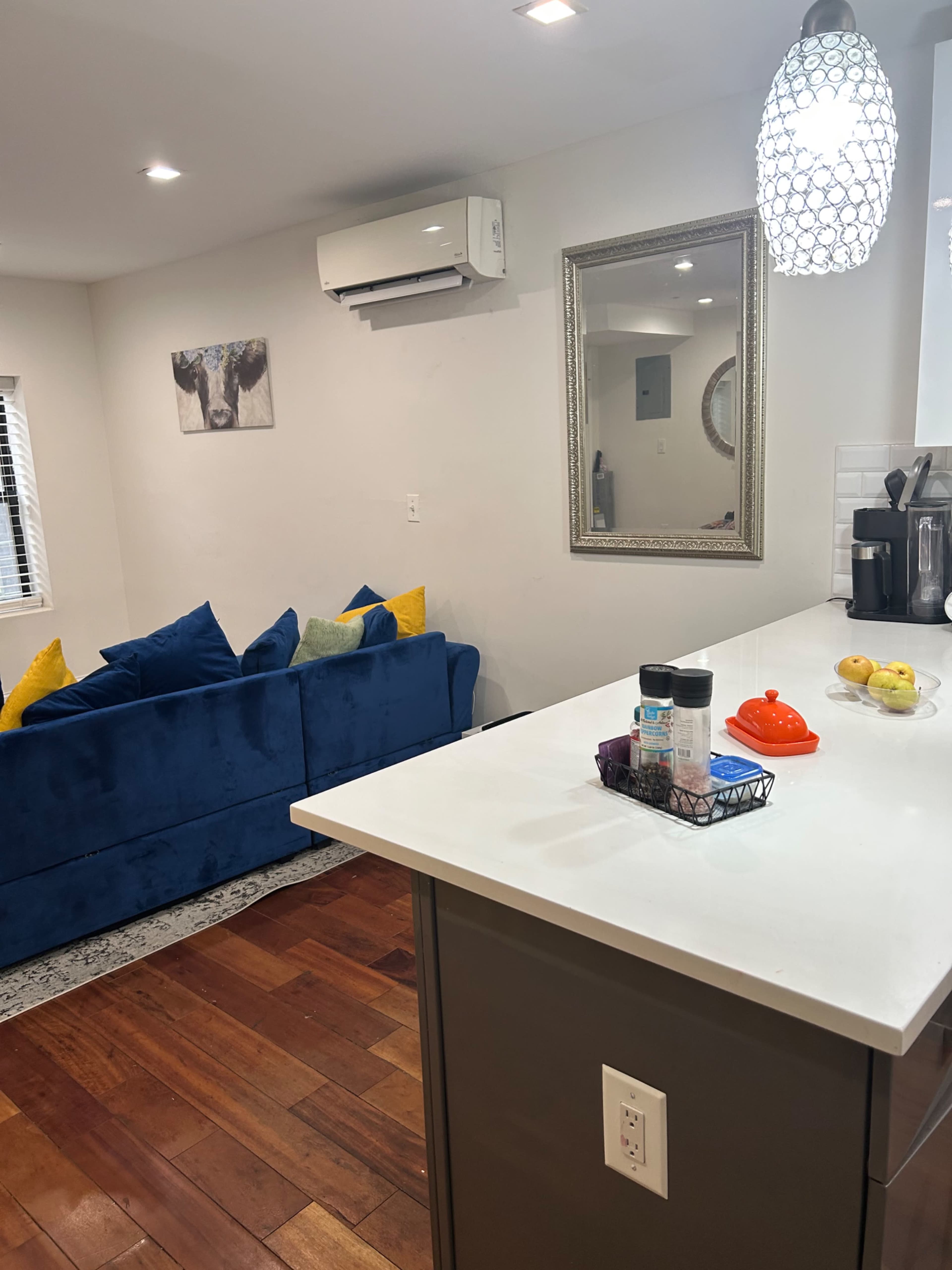 The image shows a modern living space with a blue couch adorned with yellow pillows, a kitchen island with various kitchen items, and a mirror on the wall opposite the couch.