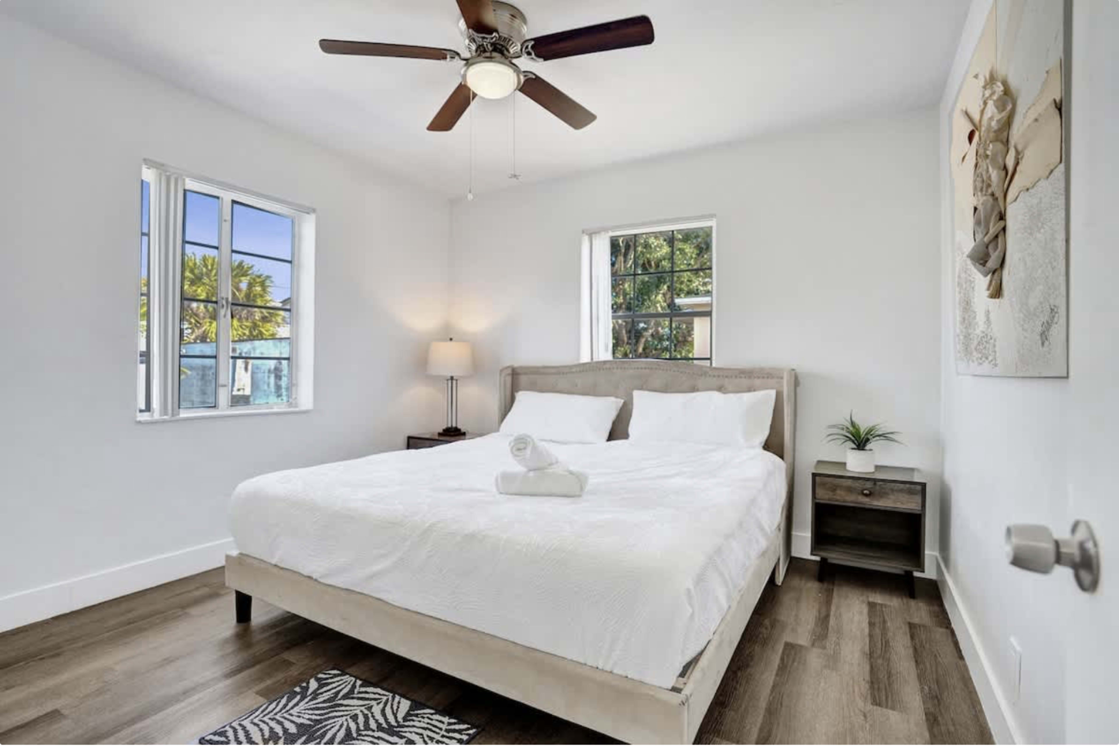 The image shows a simple bedroom featuring a large bed with white linens, a ceiling fan, two windows, and a small bedside table.