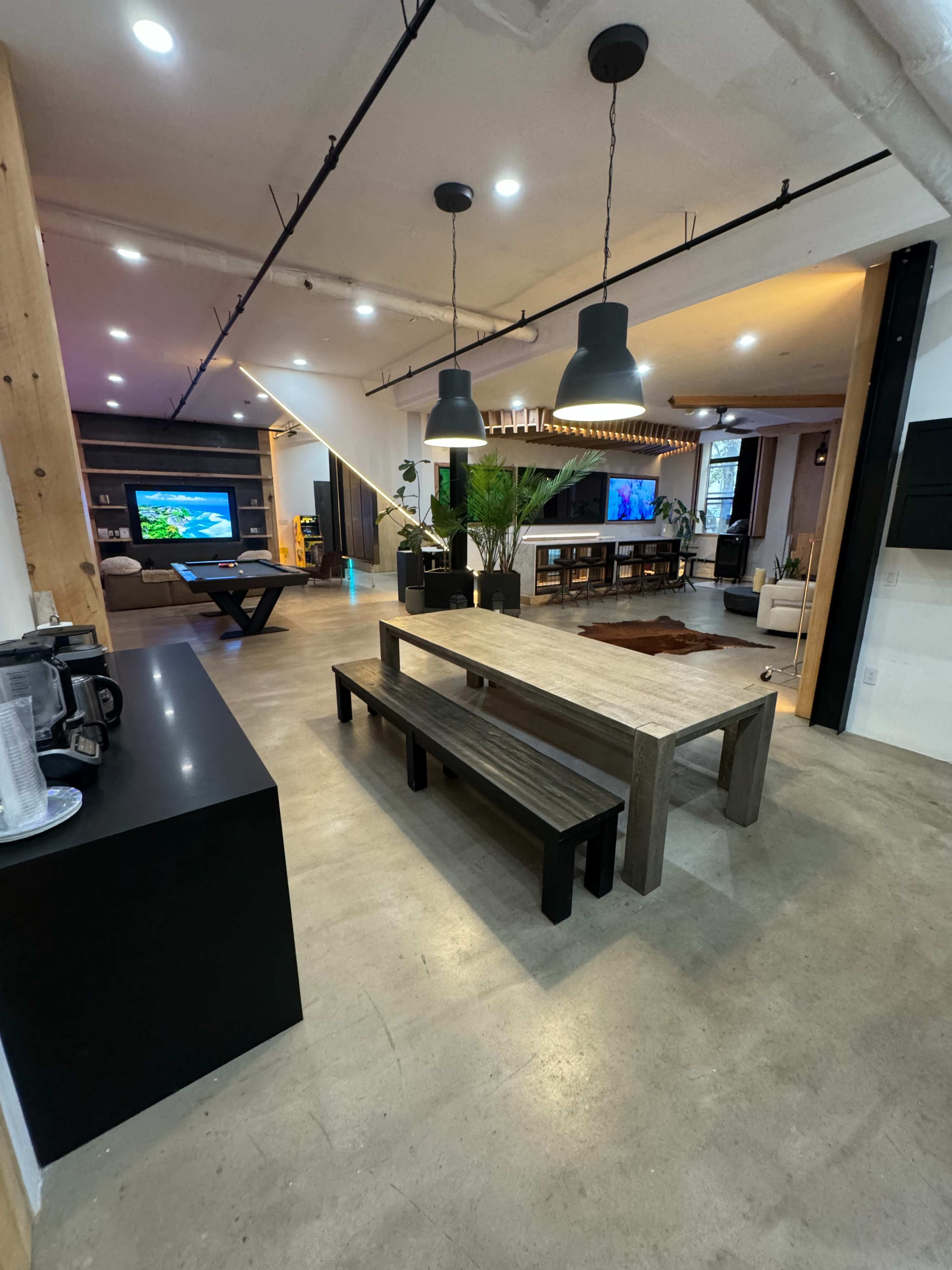 The image shows a modern open-plan living space with a large dining table, a kitchen area, a seating area, and a television in view.