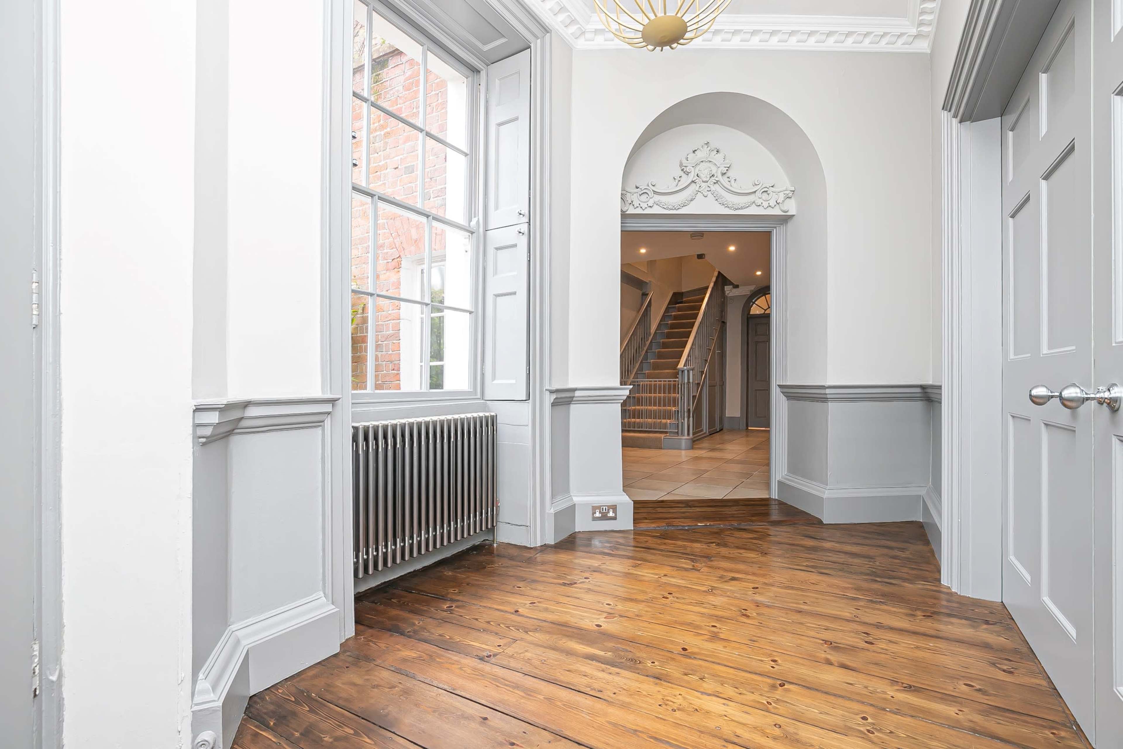 A hallway with wooden flooring, grey walls, large windows, and an archway leading to a staircase.