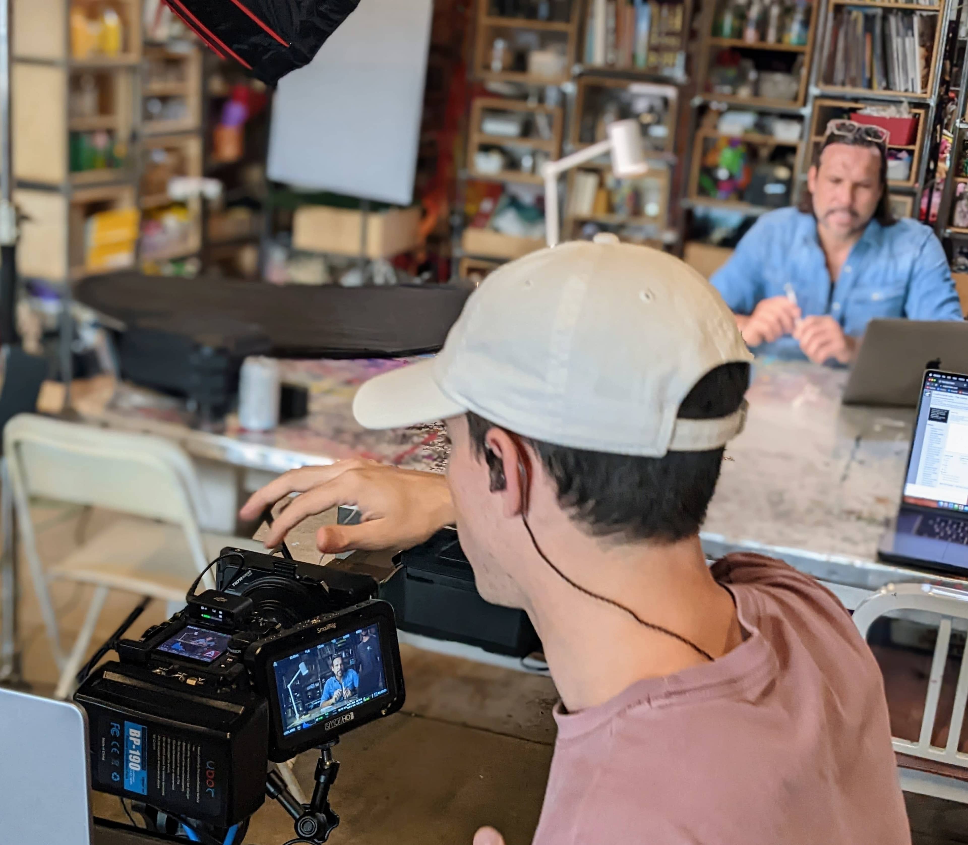 A person is filming another individual seated at a table in a cluttered workspace, using a camera mounted on a tripod.