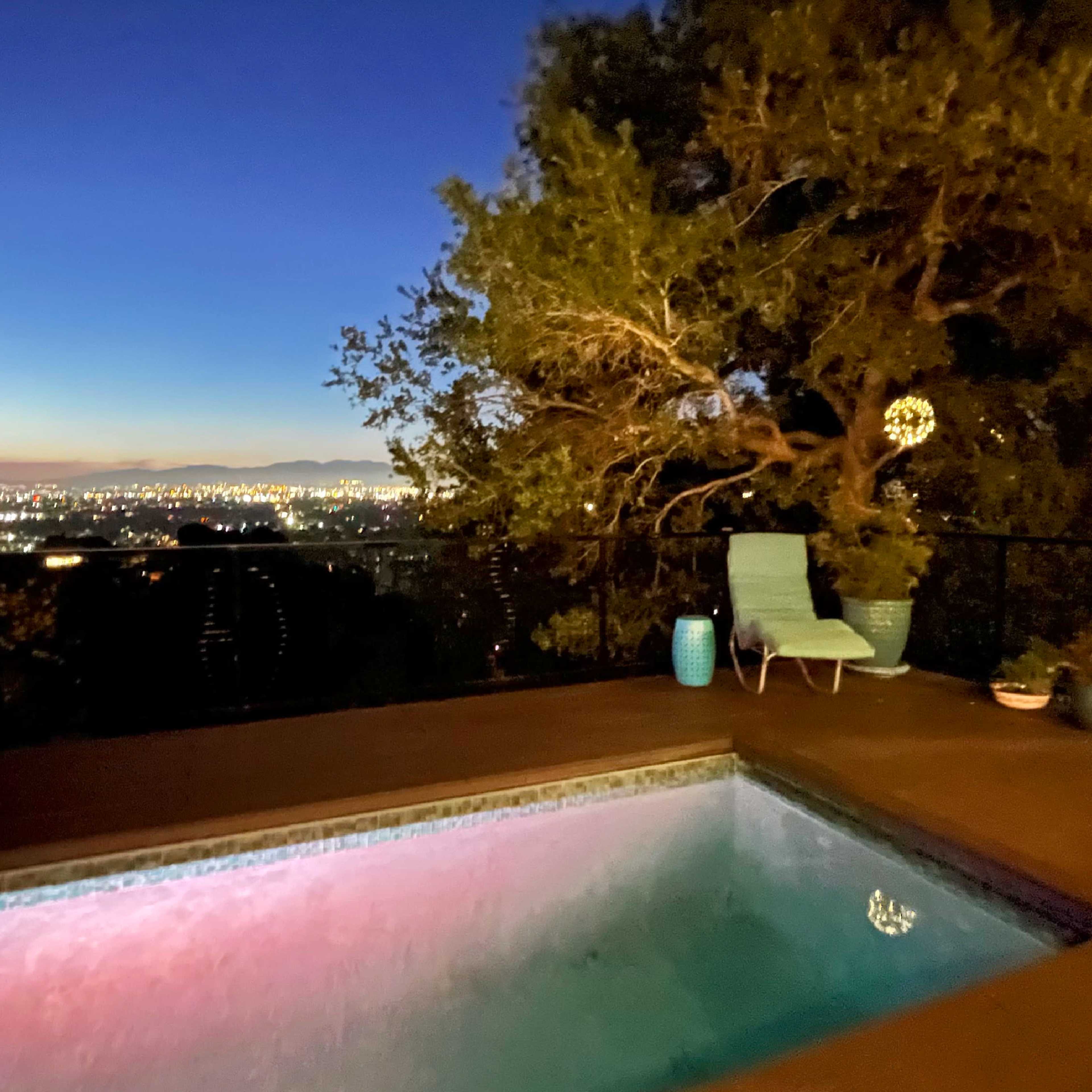 A swimming pool with illuminated water is positioned on a terrace overlooking a cityscape at dusk.