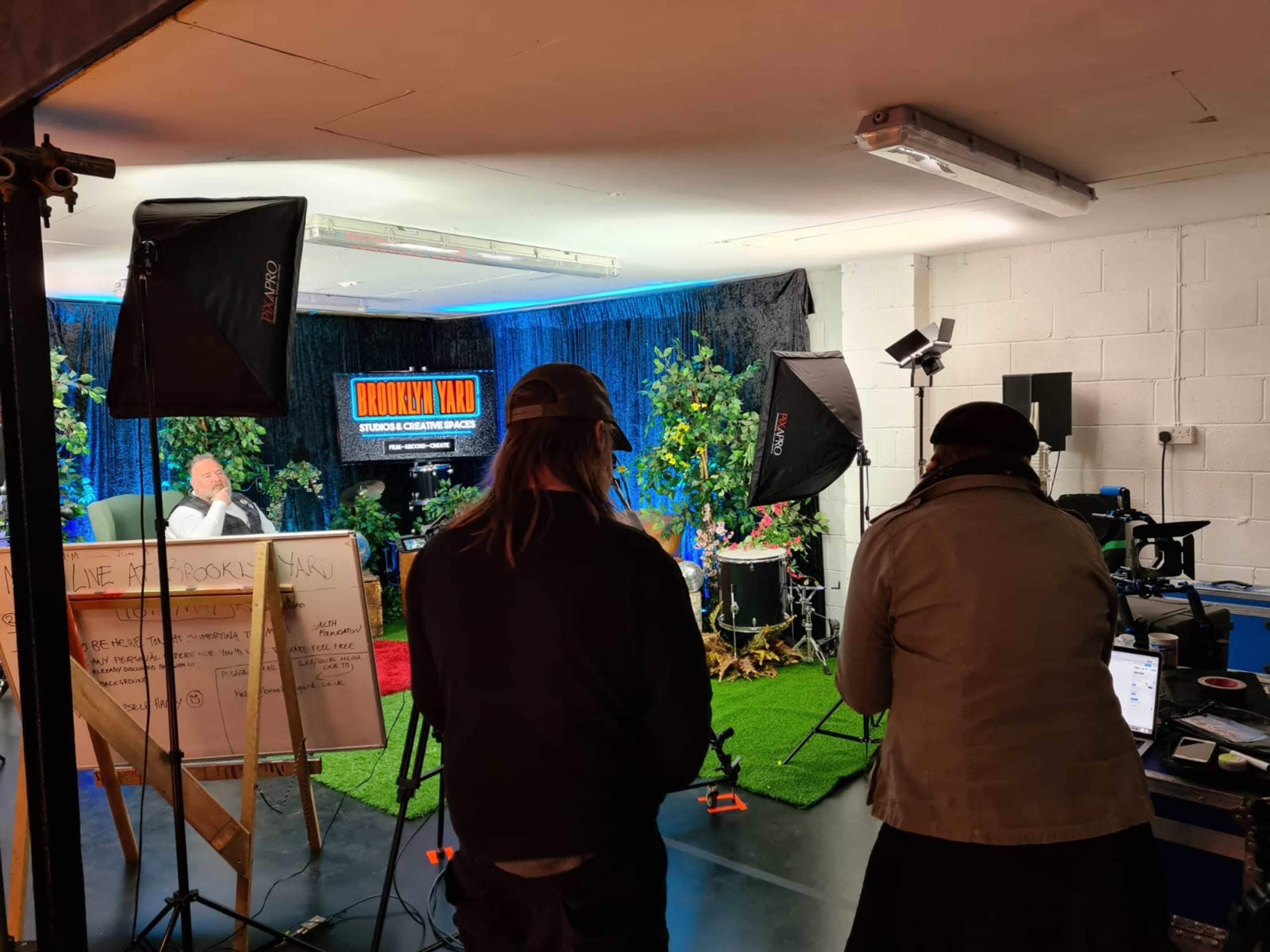 A film crew sets up equipment in a studio adorned with greenery for a live broadcast.