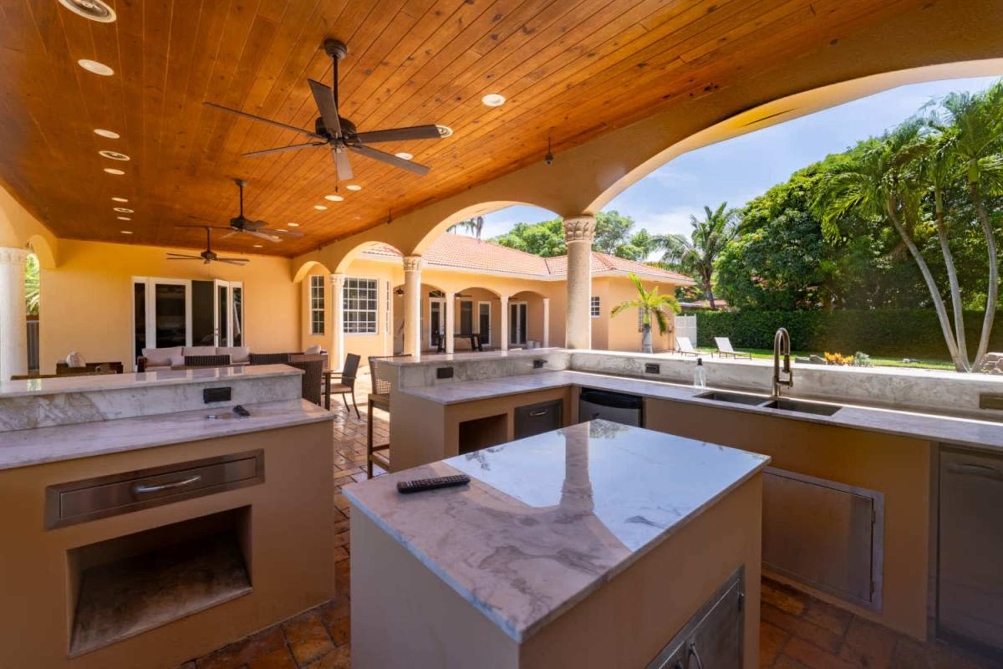 The image shows a spacious outdoor kitchen featuring marble countertops, two ceiling fans, and arched ceilings, with a view of a landscaped backyard.