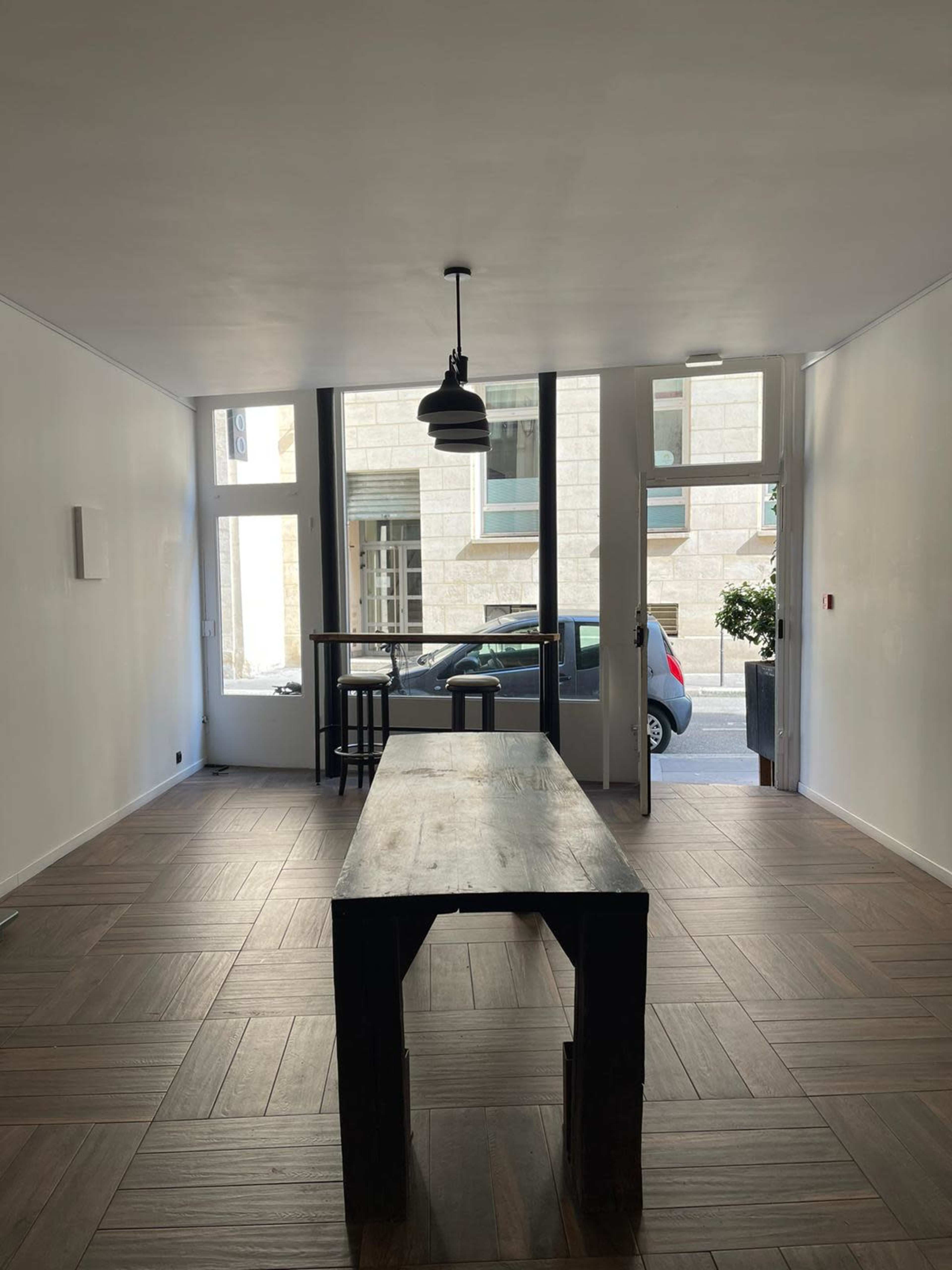 A spacious interior with a large wooden table in the center, flanked by tall windows and a doorway leading to the street.