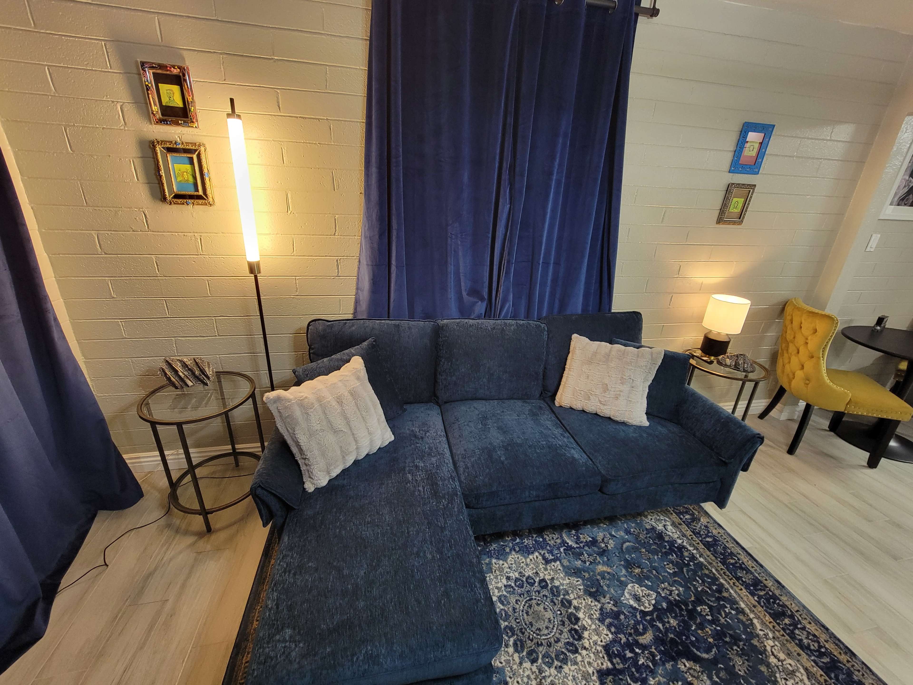 The image shows a living room with a dark blue sectional sofa, a standing lamp, and a round table with a yellow chair, set against a backdrop of blue curtains and framed artwork on the walls.