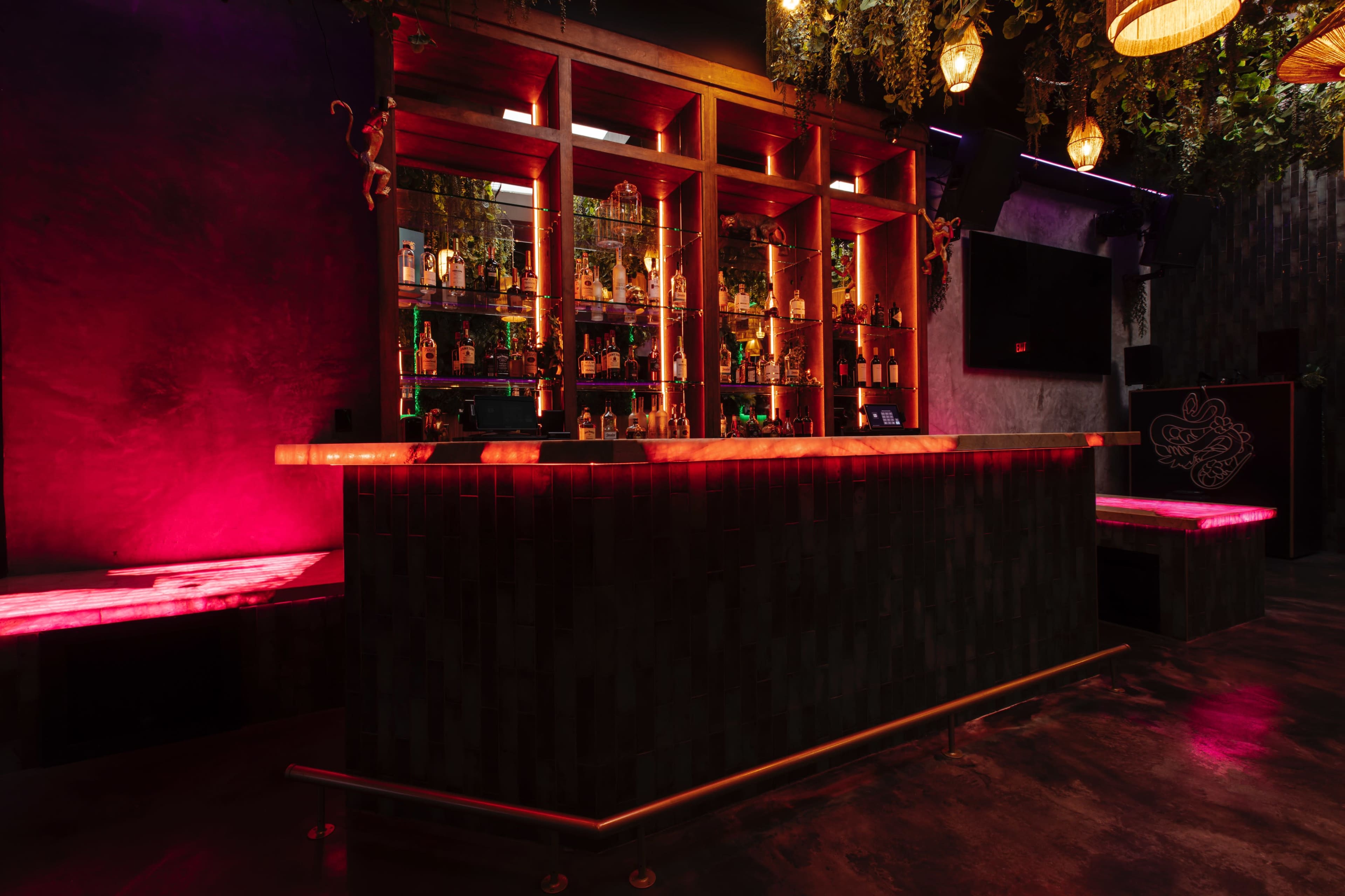 A modern bar illuminated with colorful lighting, featuring a neatly arranged display of liquor bottles behind a wooden counter.
