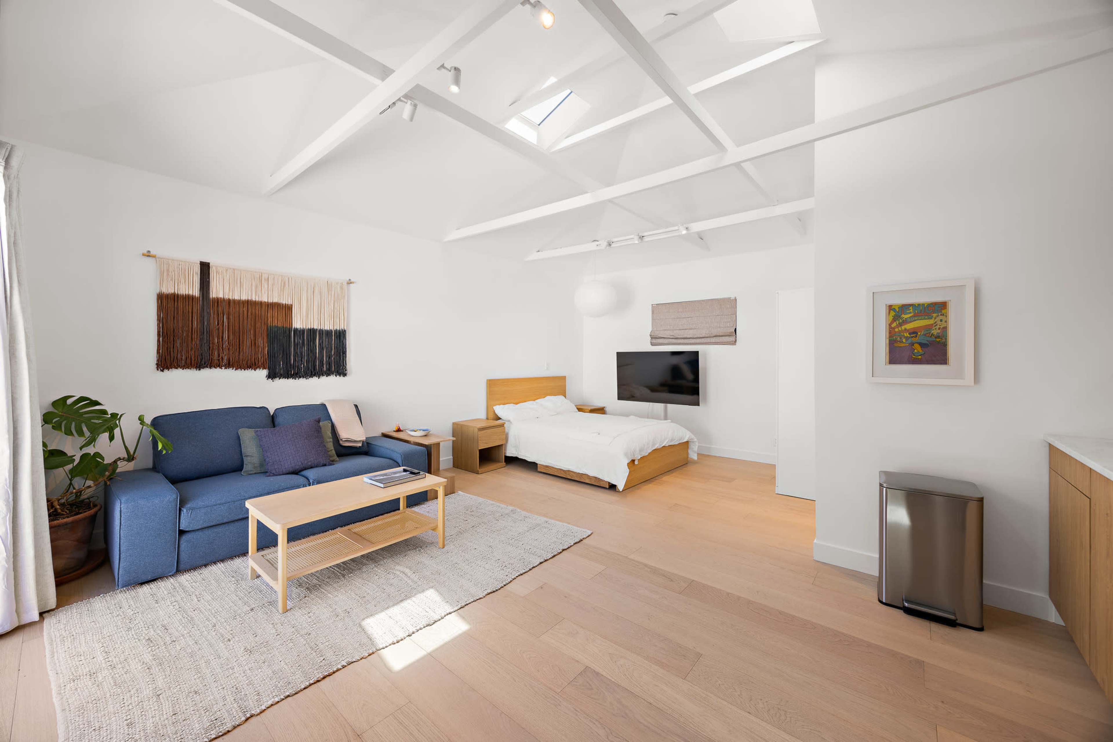 The image shows a minimalist bedroom with a blue sofa, a wooden bed, a television mounted on the wall, and a large window allowing natural light to enter.