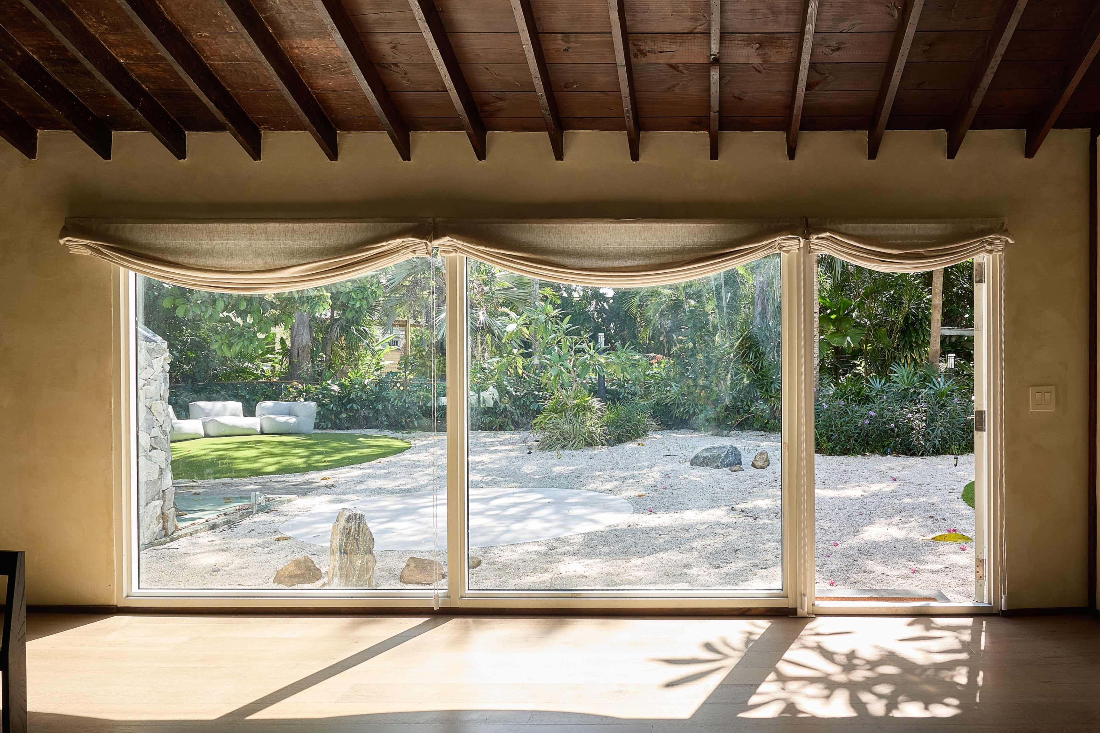 The image features a large sliding glass door with draped curtains, leading to a landscaped garden with a circular patio area and lush greenery.