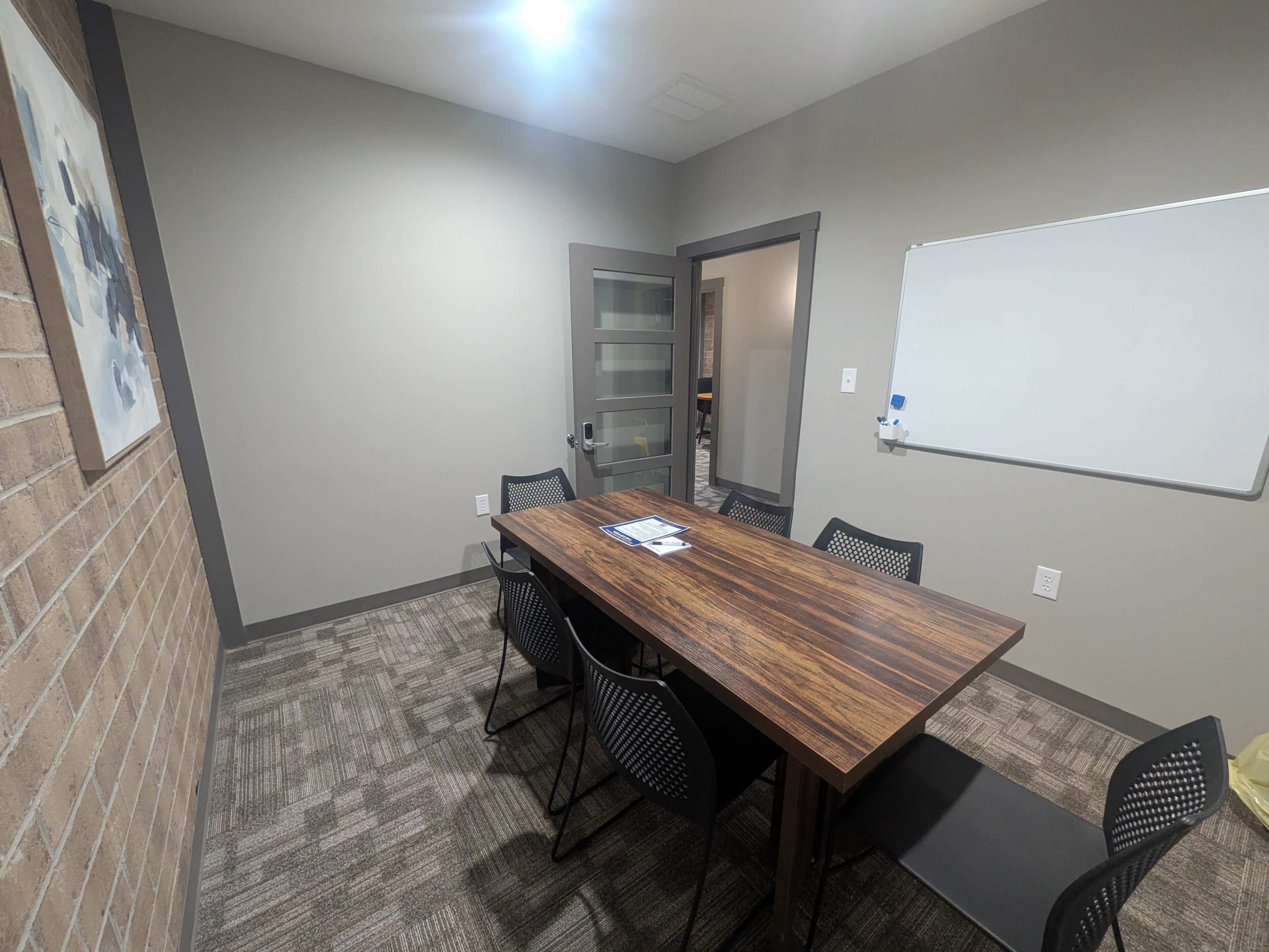 The image shows a conference room with a wooden table surrounded by six black chairs, a whiteboard on the wall, and a glass door leading to another room.