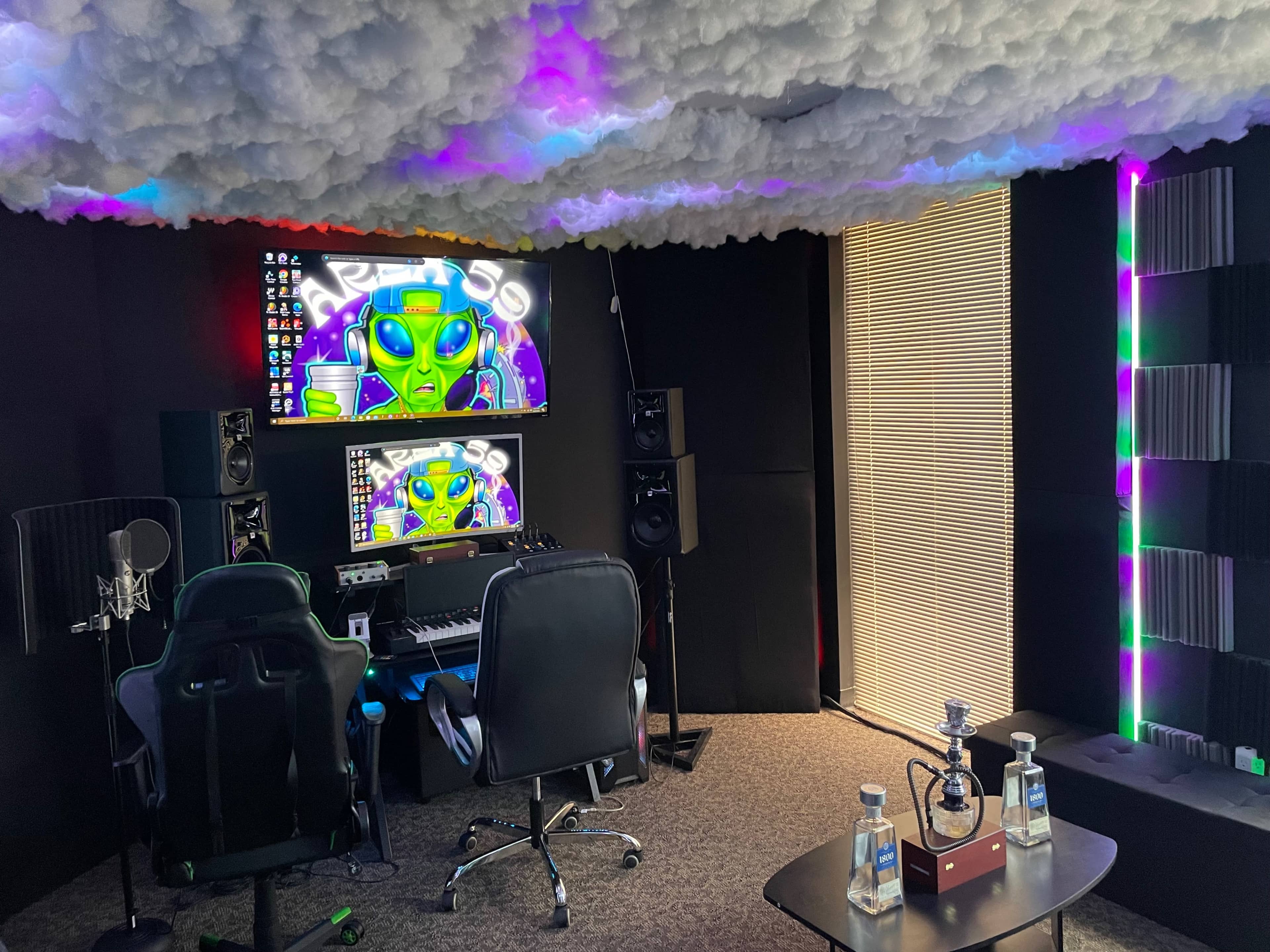 A music studio featuring a desk with a digital audio workstation, two monitors displaying colorful graphics, a microphone setup, and colorful lighting against a textured ceiling.