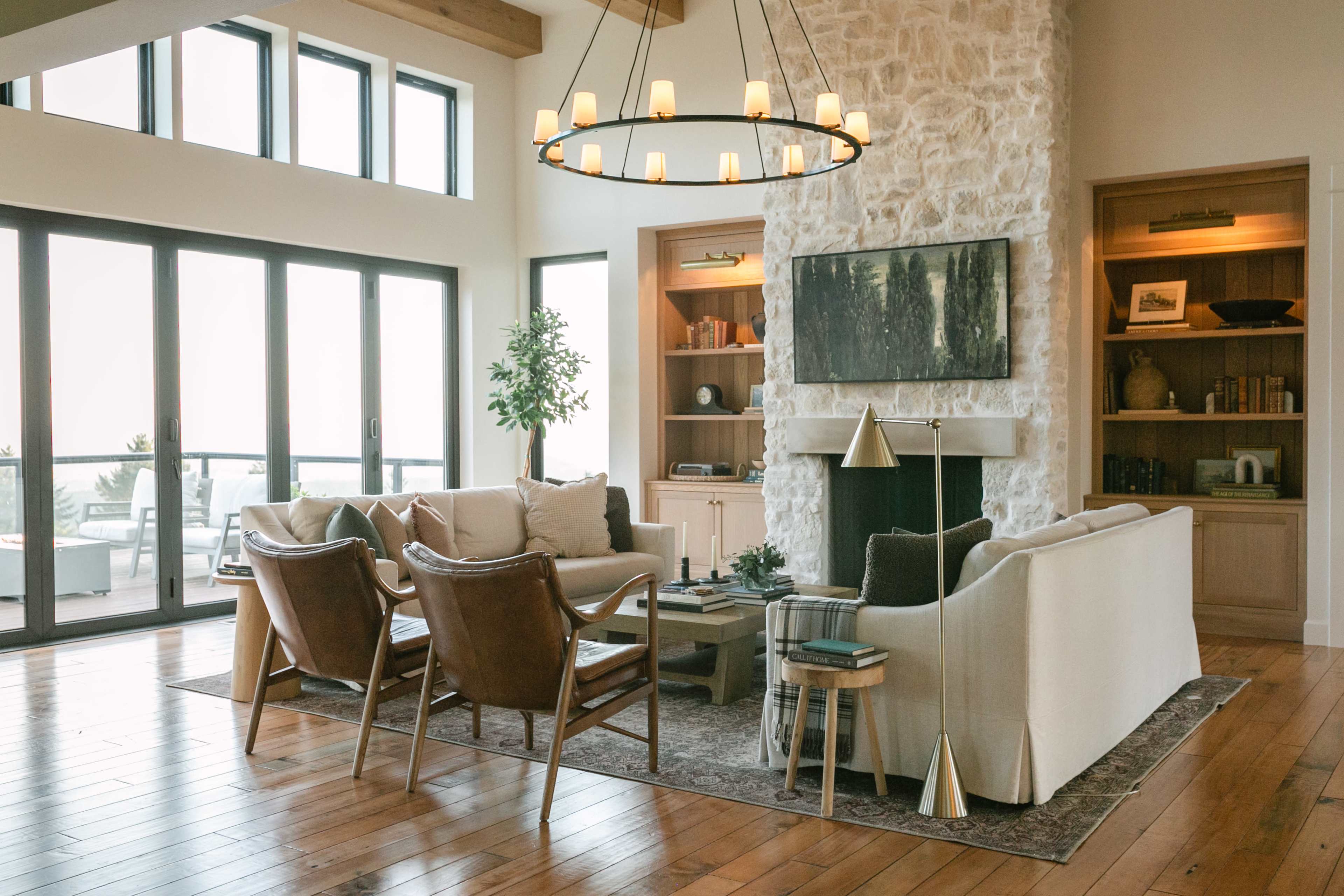 A spacious living room features a stone wall, large windows, a chandelier, and modern furniture arranged around a coffee table.