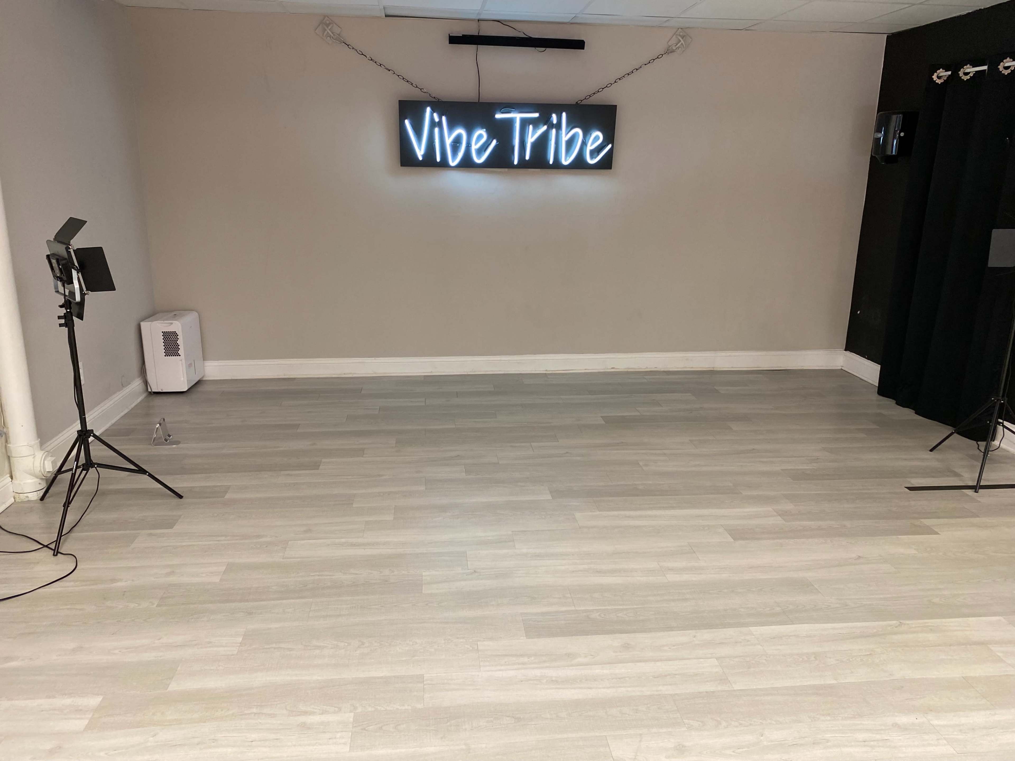 An empty room with light-colored wooden flooring and a neon sign reading "Vibe Tribe" on the wall.