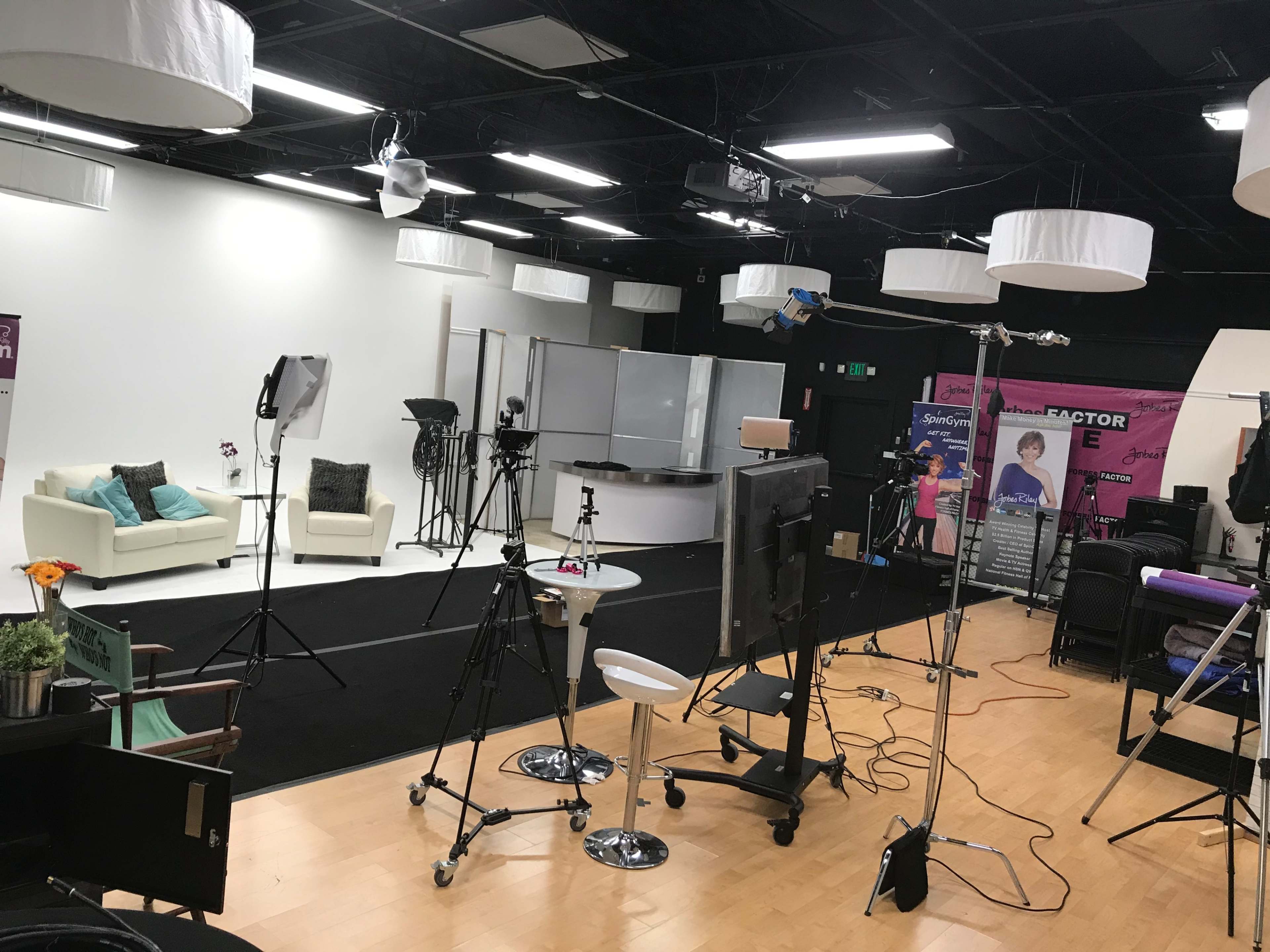A studio set up for a production, featuring a seating area, multiple cameras, and lighting equipment arranged on a black carpeted floor.