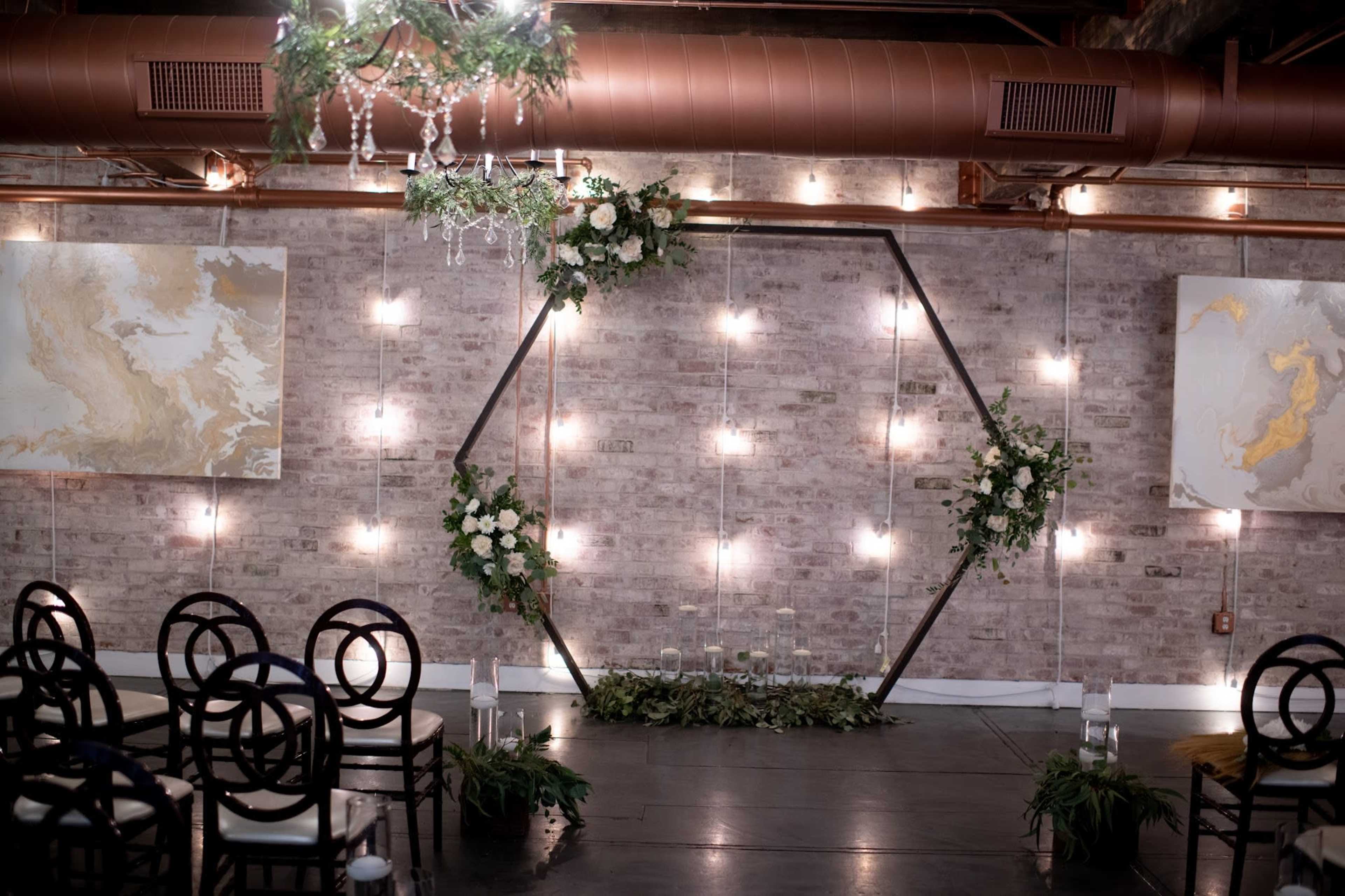 A wedding ceremony setup featuring an illuminated hexagonal arch adorned with greenery and white flowers, set against a brick wall with abstract artwork.