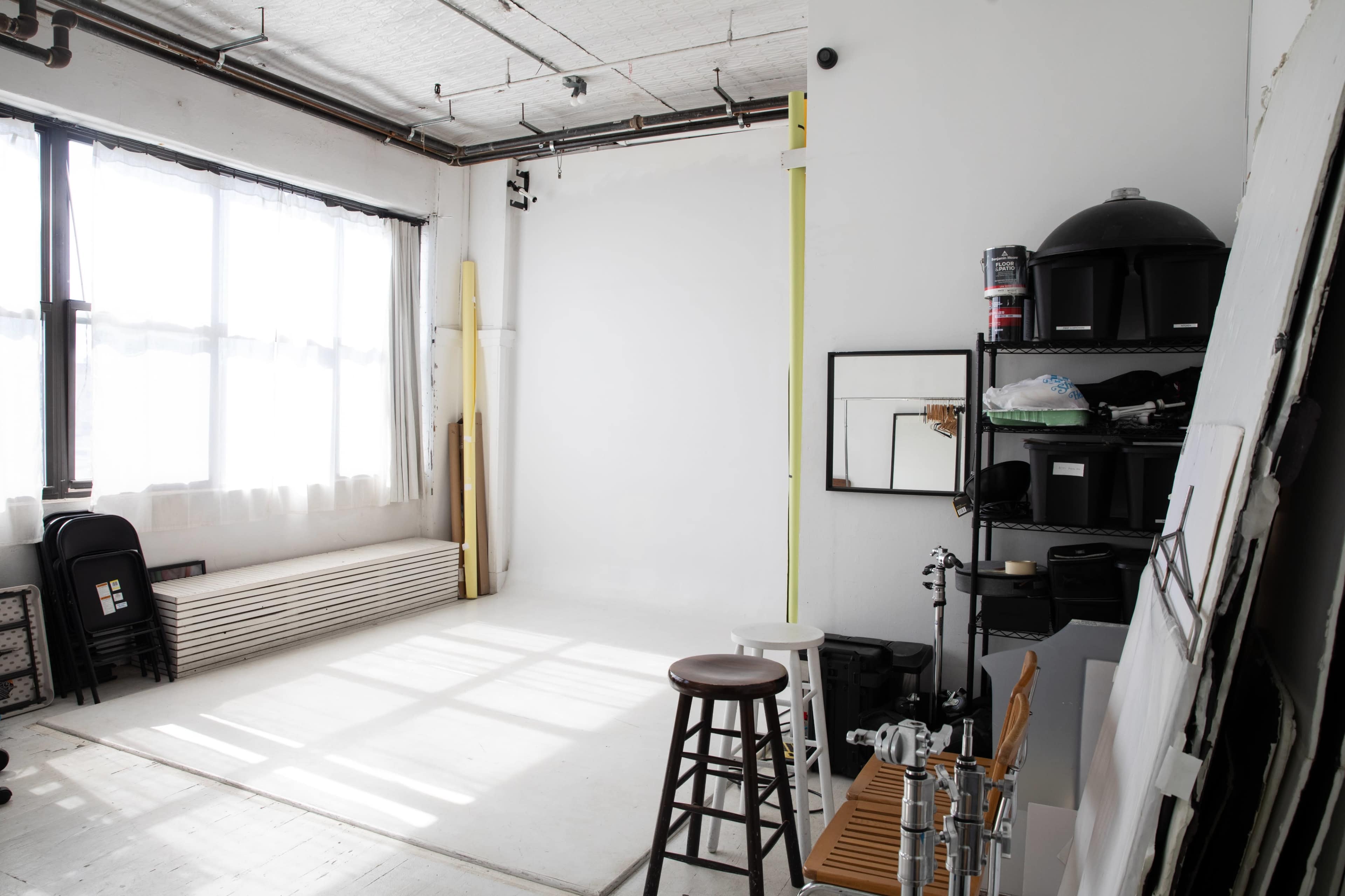 The image shows a bright, minimalist photography studio with large windows, a blank wall, and various equipment and furniture arranged neatly around the space.