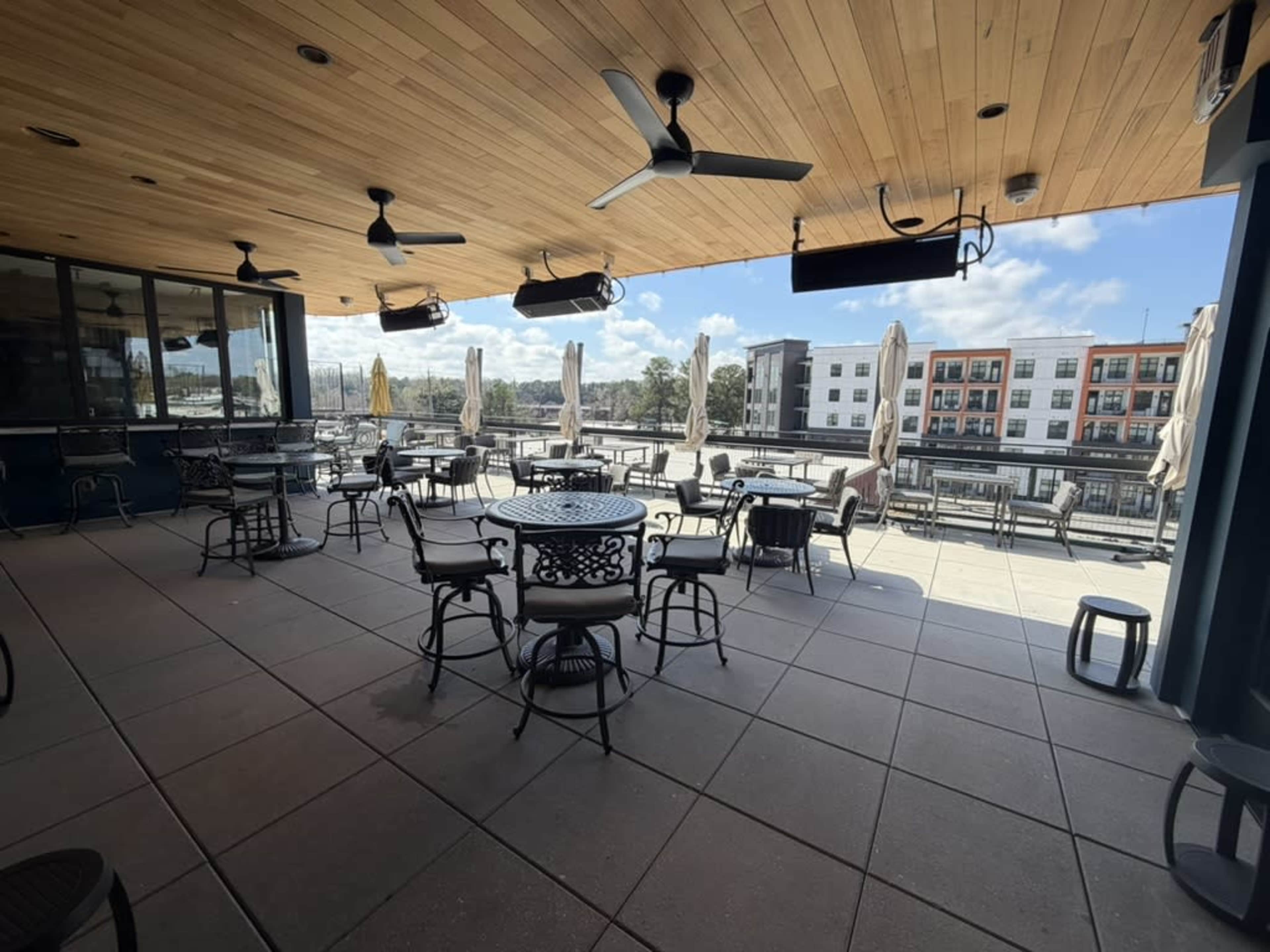 The image shows an outdoor seating area with metal tables and chairs on a patio, featuring a wooden ceiling and a view of nearby buildings and a cloudy sky.