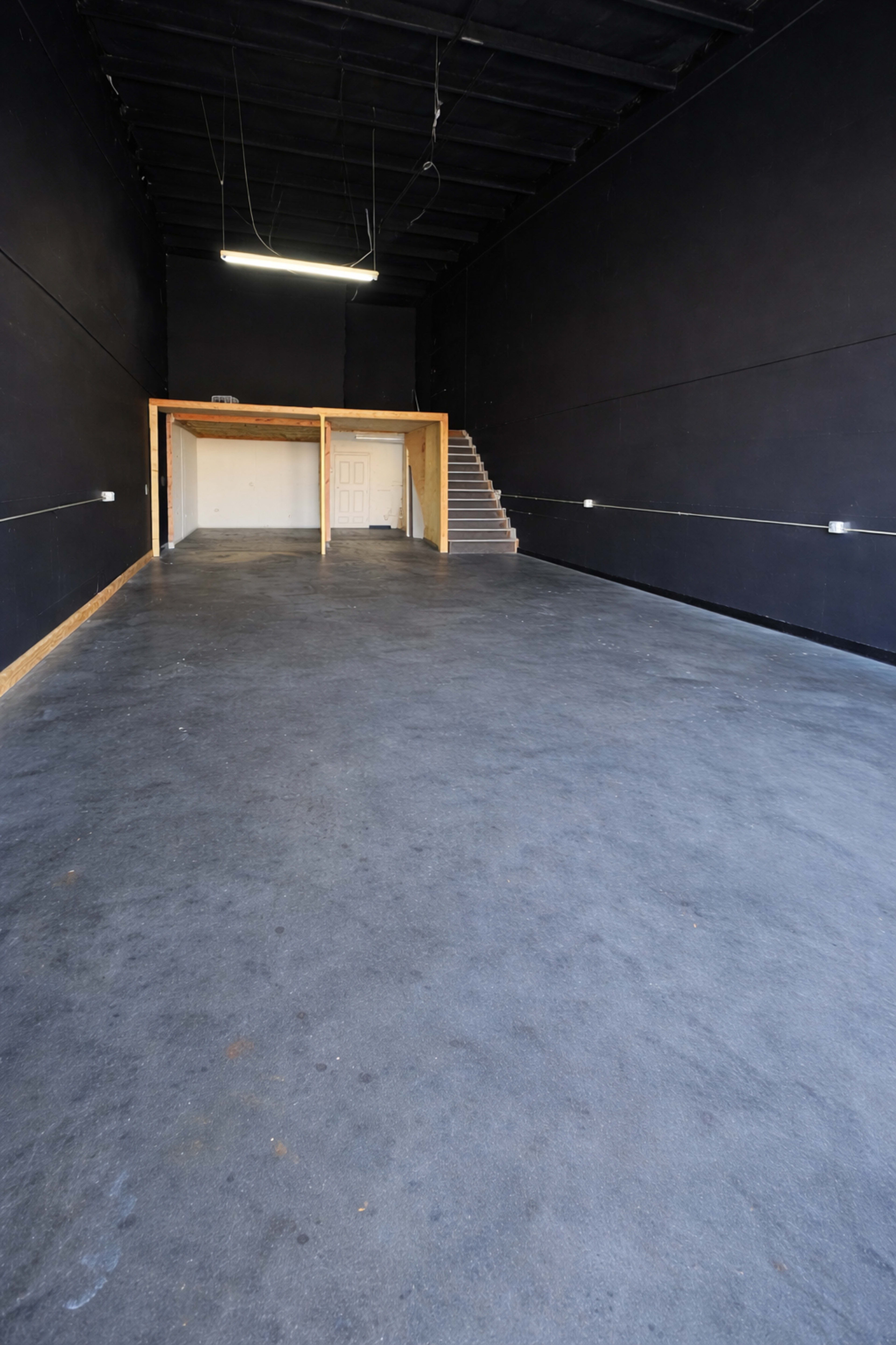 The image shows an empty industrial space with black walls, a concrete floor, a small room at the back, and a staircase on the right side.