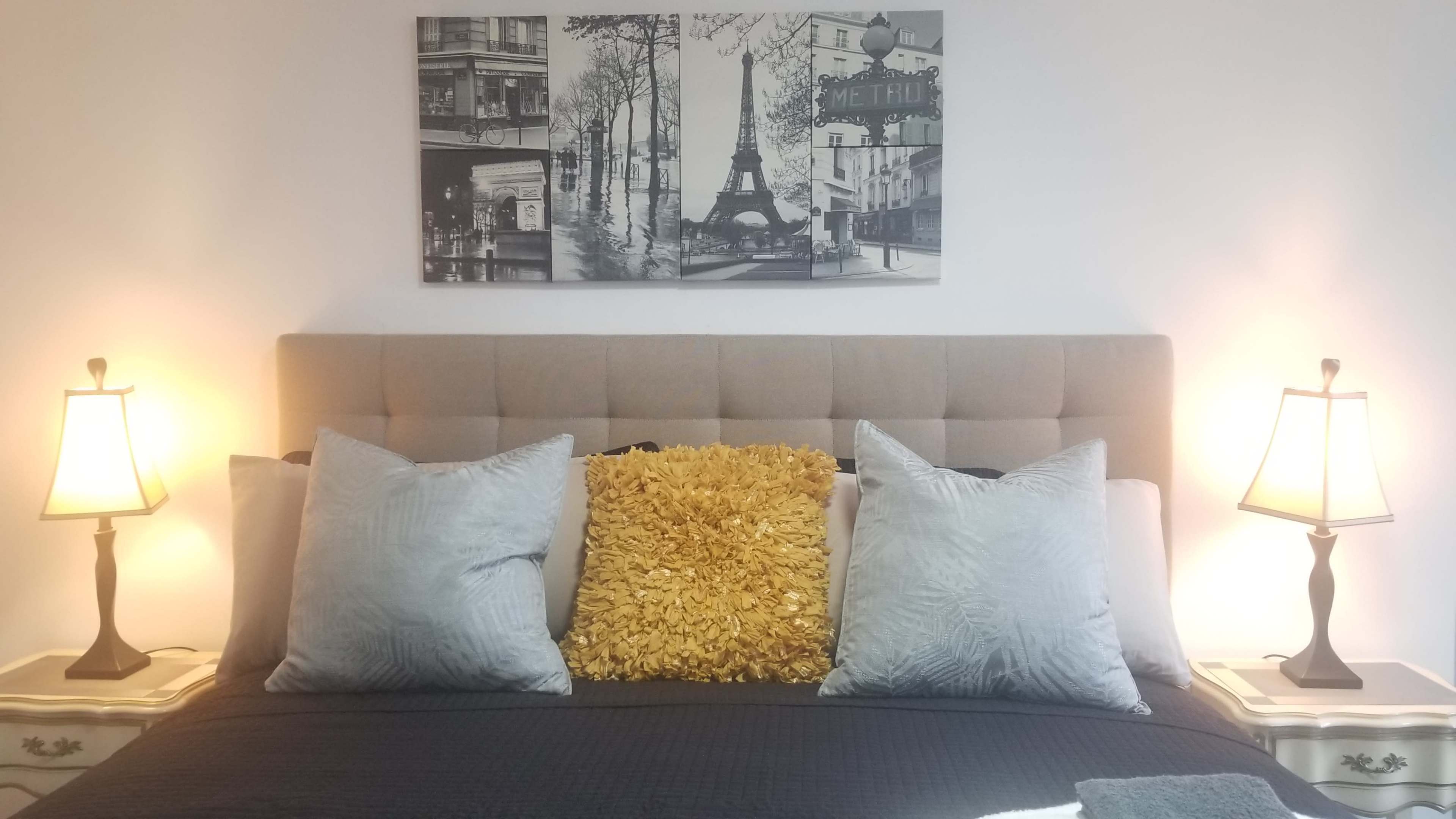 A neatly made bed with gray pillows and a yellow decorative pillow, flanked by two lit bedside lamps, and a black and white wall art featuring city scenes.