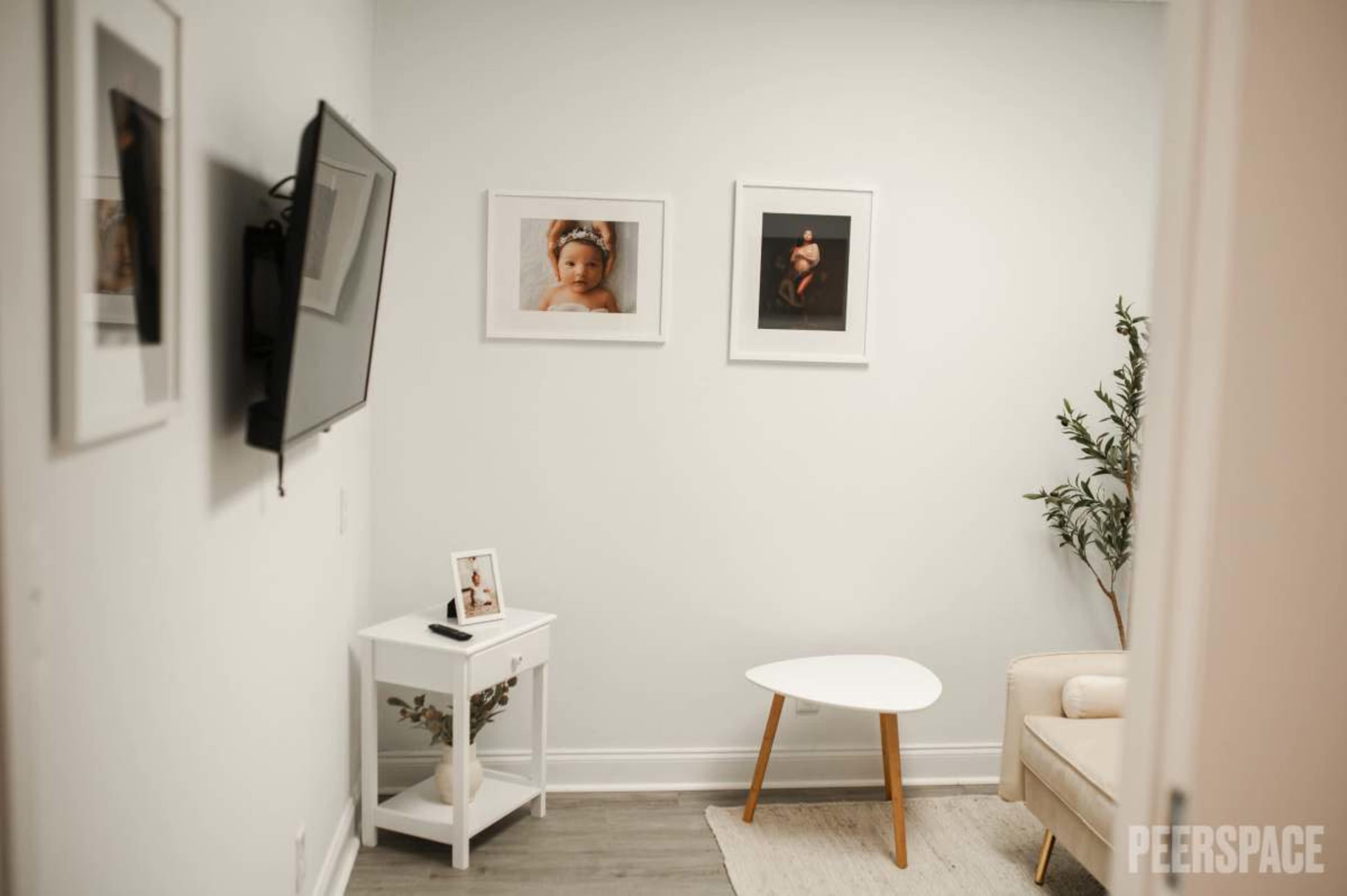 The image shows a small, modern room featuring a television mounted on the wall, two framed photographs, a white side table, a light-colored couch, and a plant.