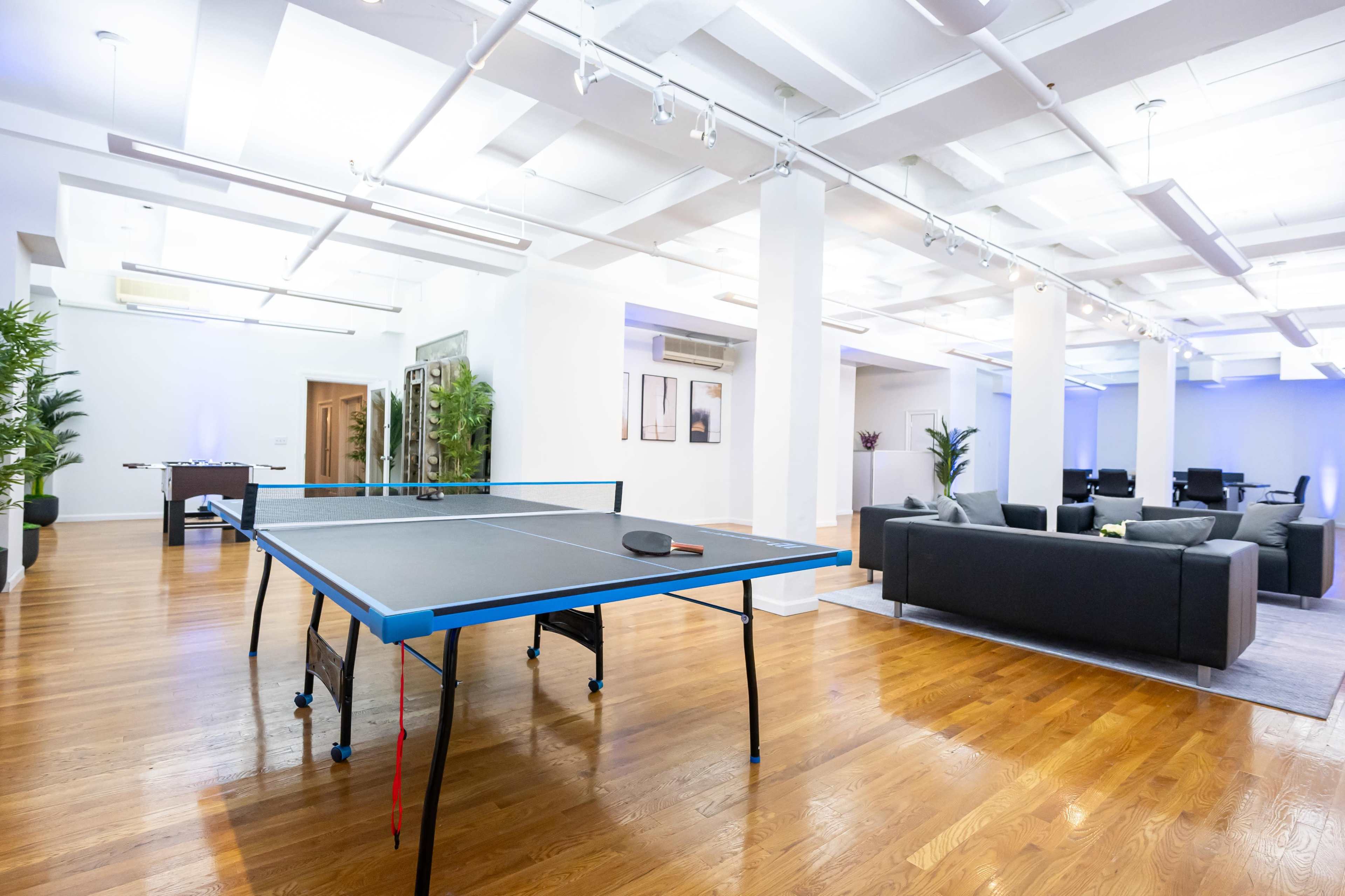 The image shows a modern, open space featuring a ping pong table in the foreground and a seating area with a couch in the background.
