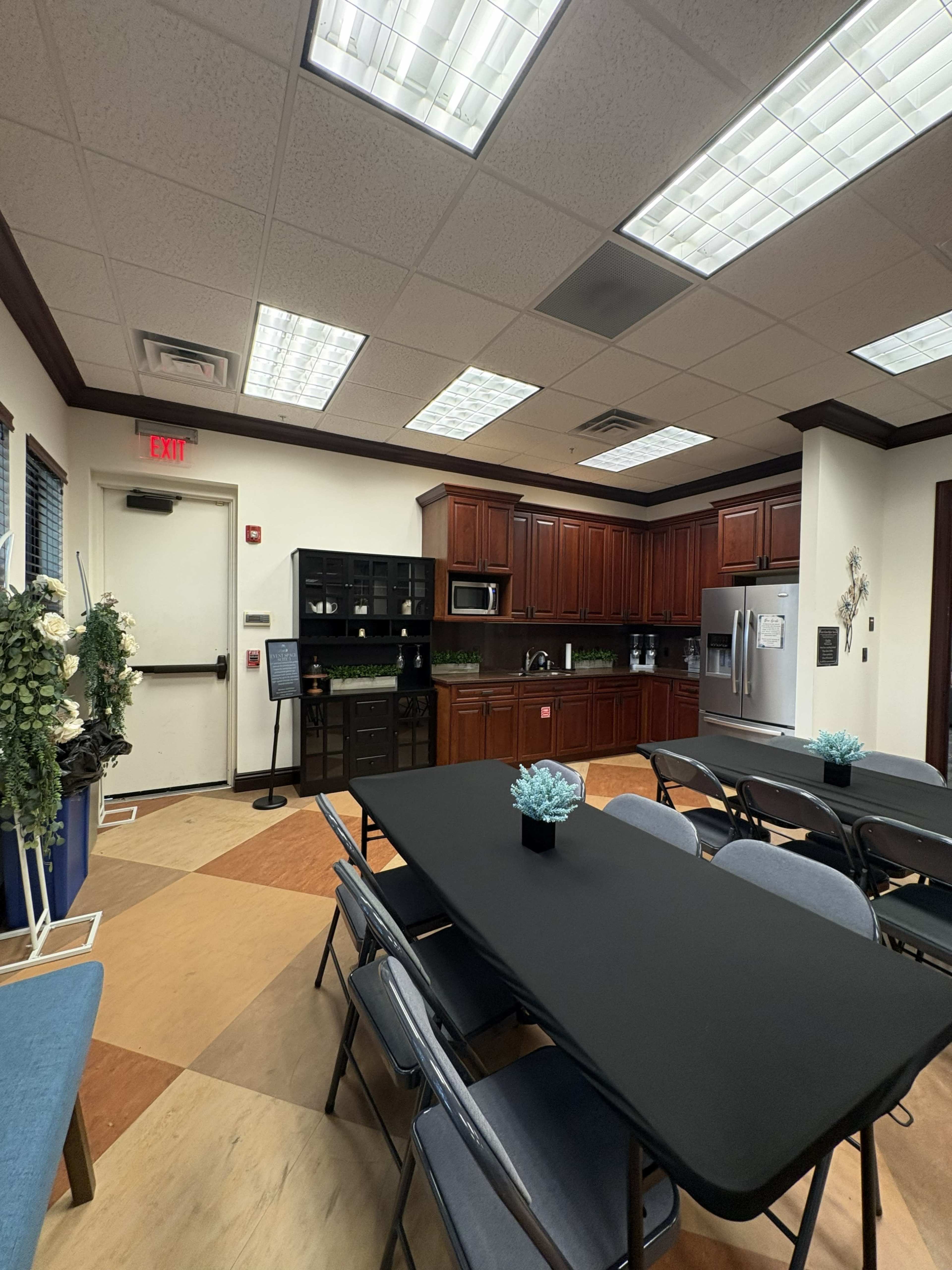 The image shows a room with a kitchenette in the corner, featuring dark wooden cabinets and stainless steel appliances, alongside a long black table surrounded by chairs.