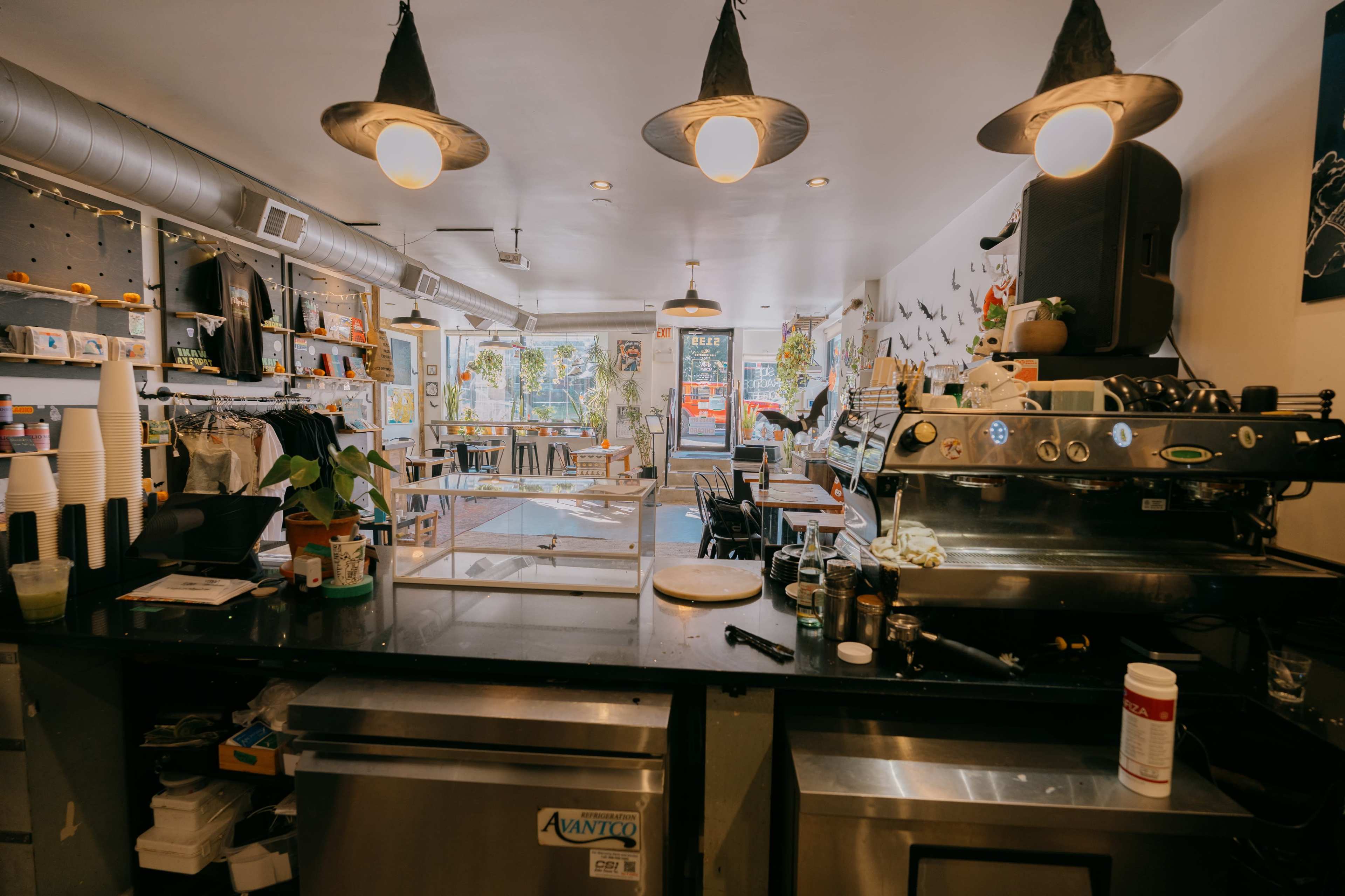 The image shows a cozy café interior with a coffee machine on the counter, tables and chairs arranged for customers, and large windows letting in natural light.