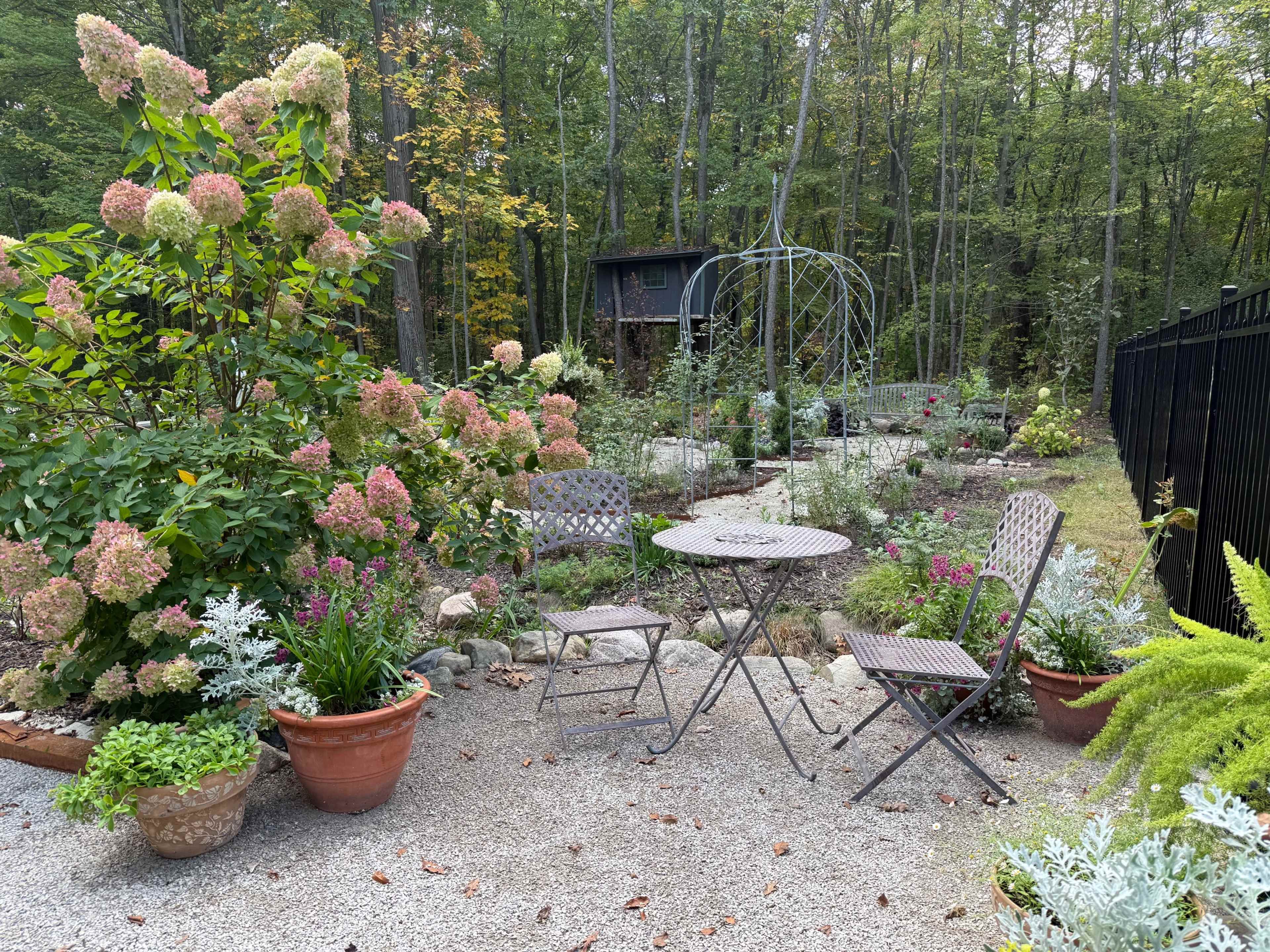 A small garden area features a bistro table and chairs surrounded by flowering plants and a wooden structure in the background.