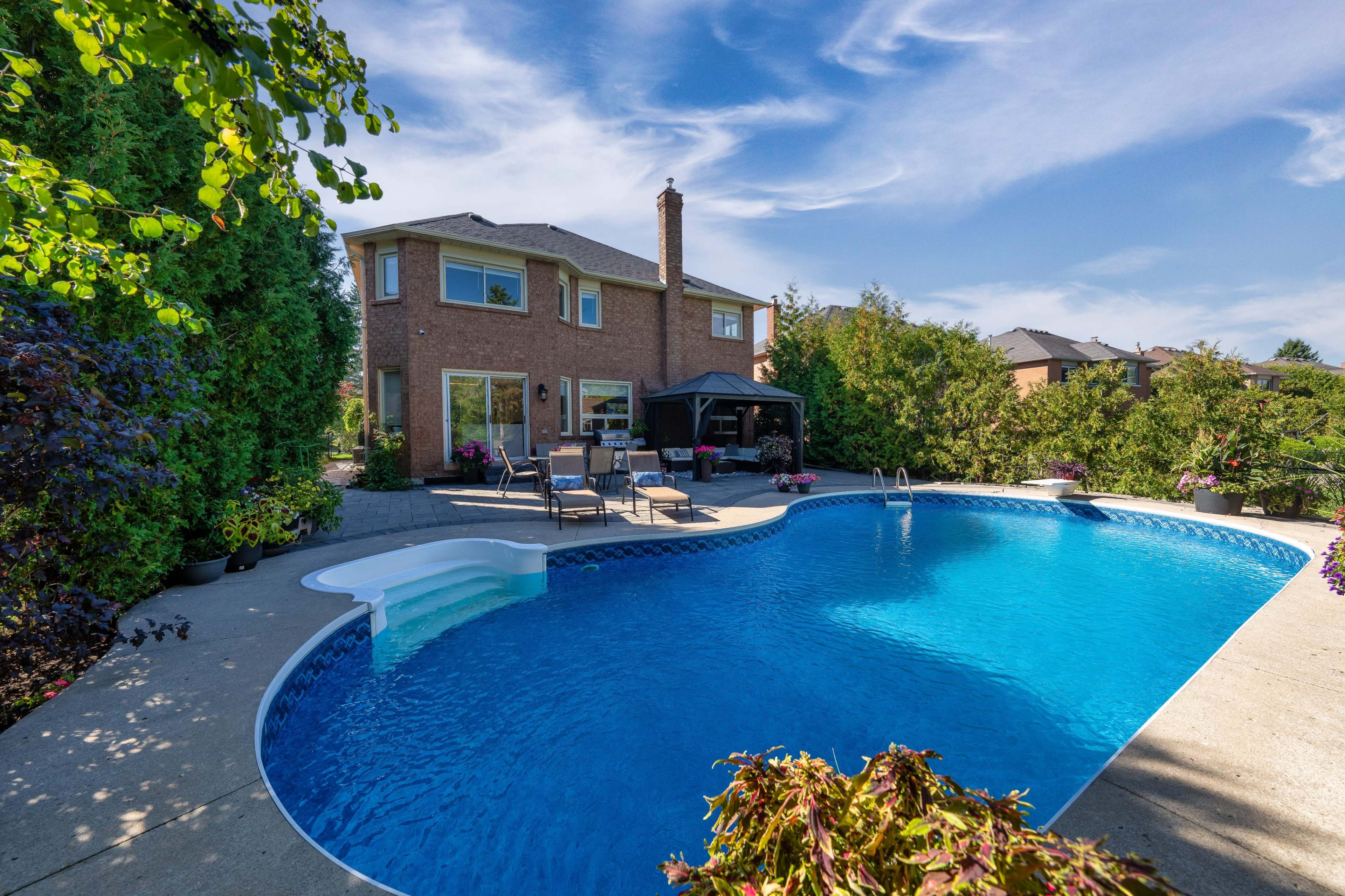 The image shows a backyard with a blue swimming pool surrounded by greenery and a two-story house in the background.