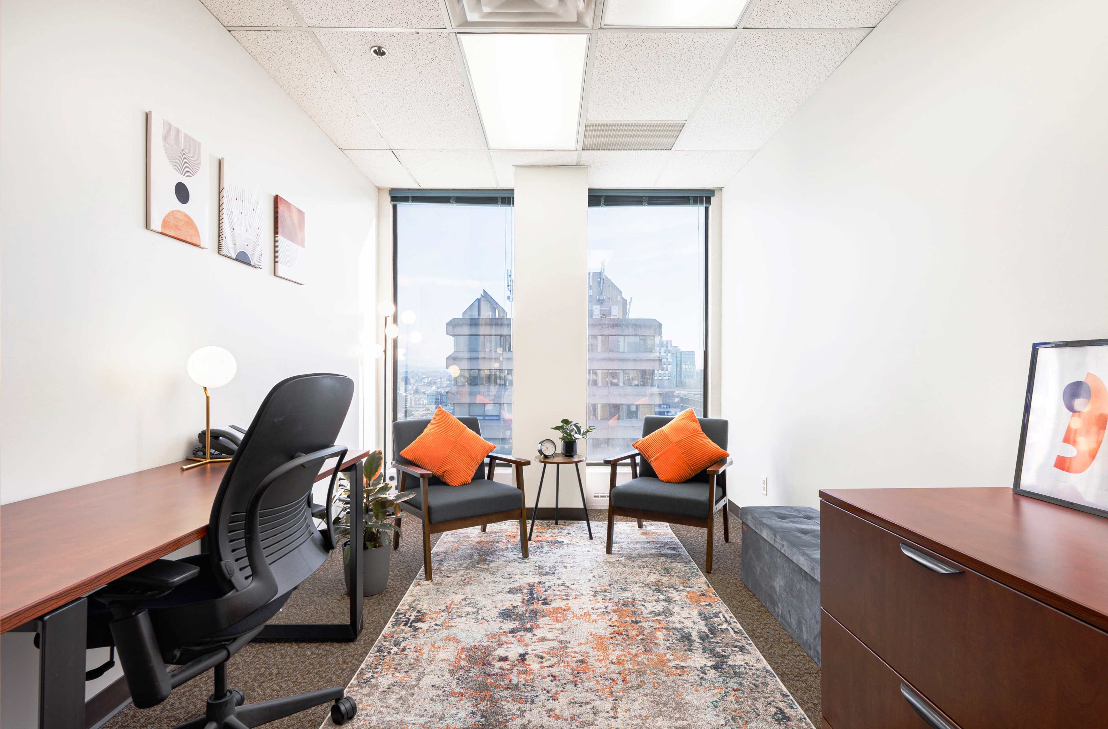 A small office with two chairs and a table, featuring a desk and a window that overlooks a cityscape.