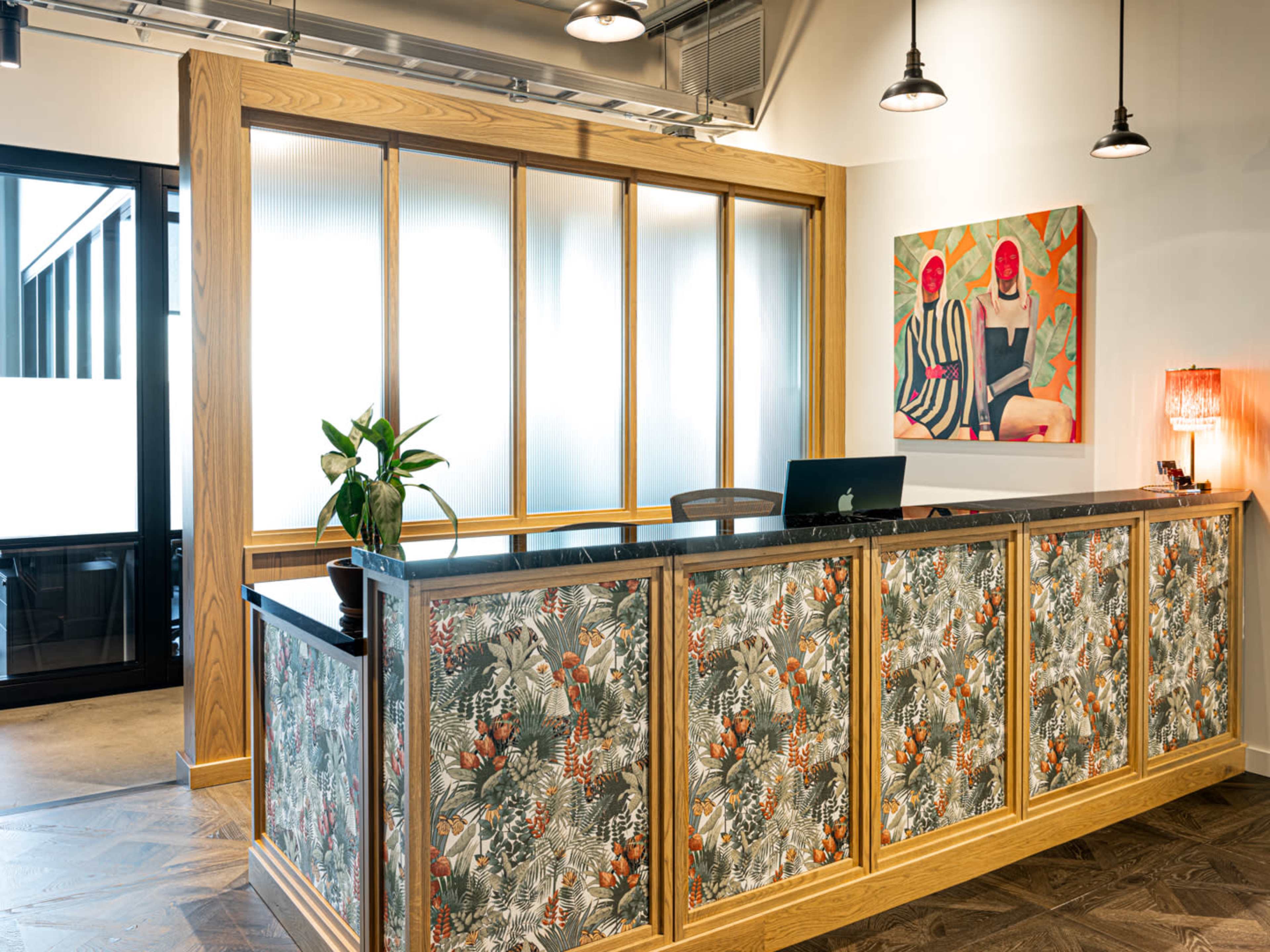 A reception desk with a floral-patterned front, a computer on the countertop, and a plant on the side, set in a modern office environment.