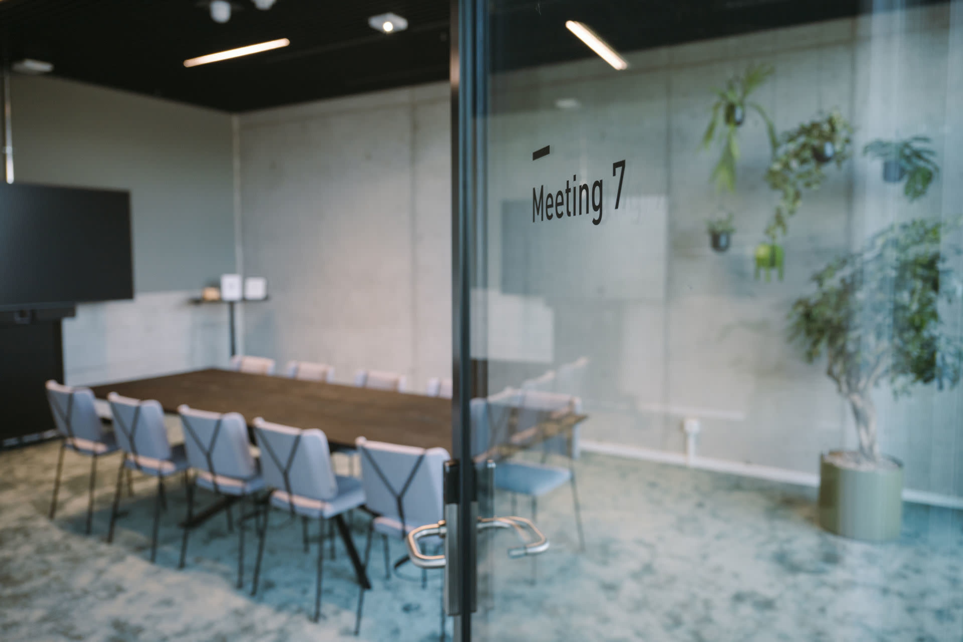 A glass door labeled "Meeting 7" opens into a conference room featuring a large table surrounded by chairs and greenery on the walls.