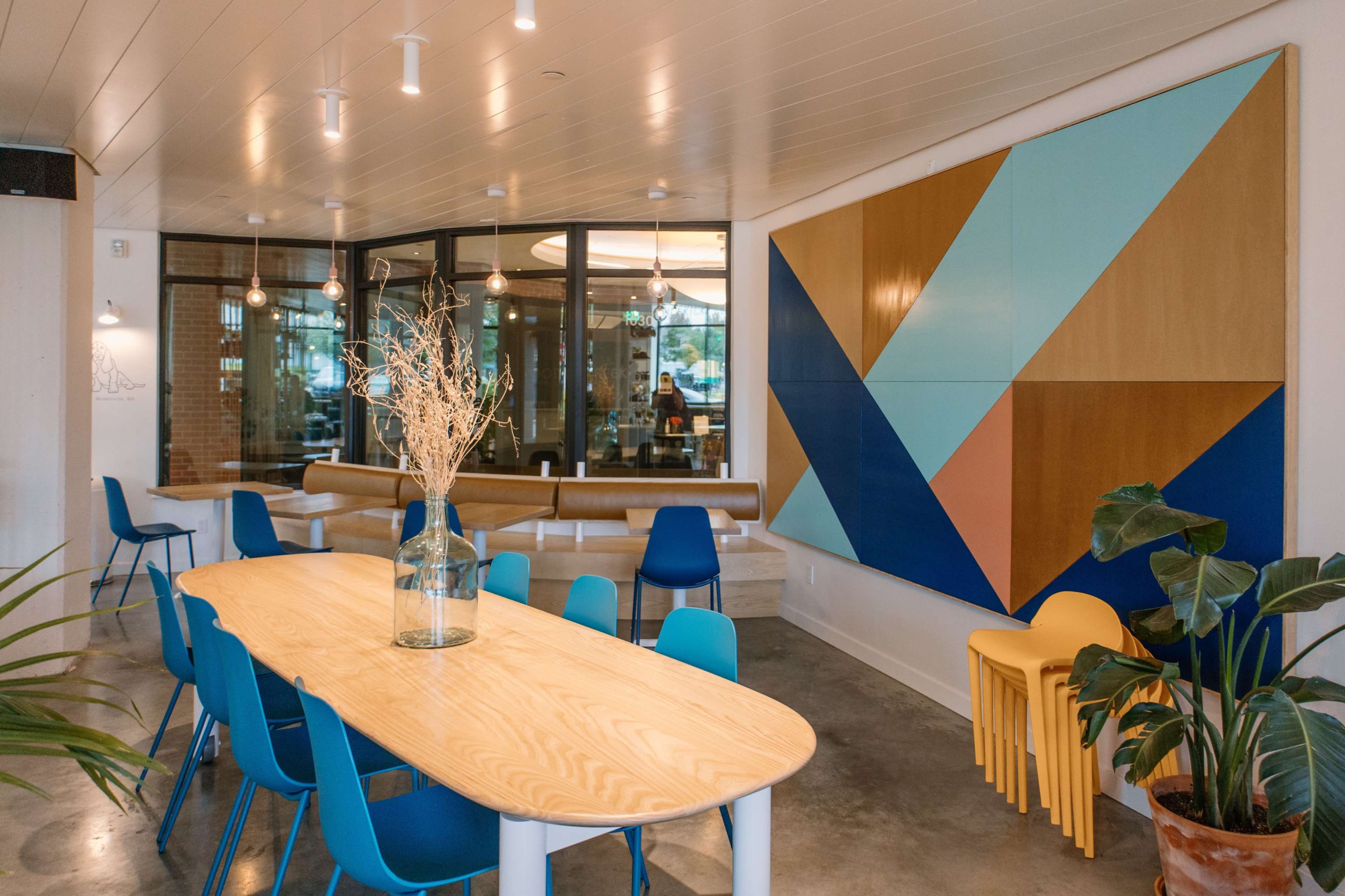 A modern interior space with a wooden table surrounded by blue chairs, large windows, and a colorful geometric artwork on the wall.