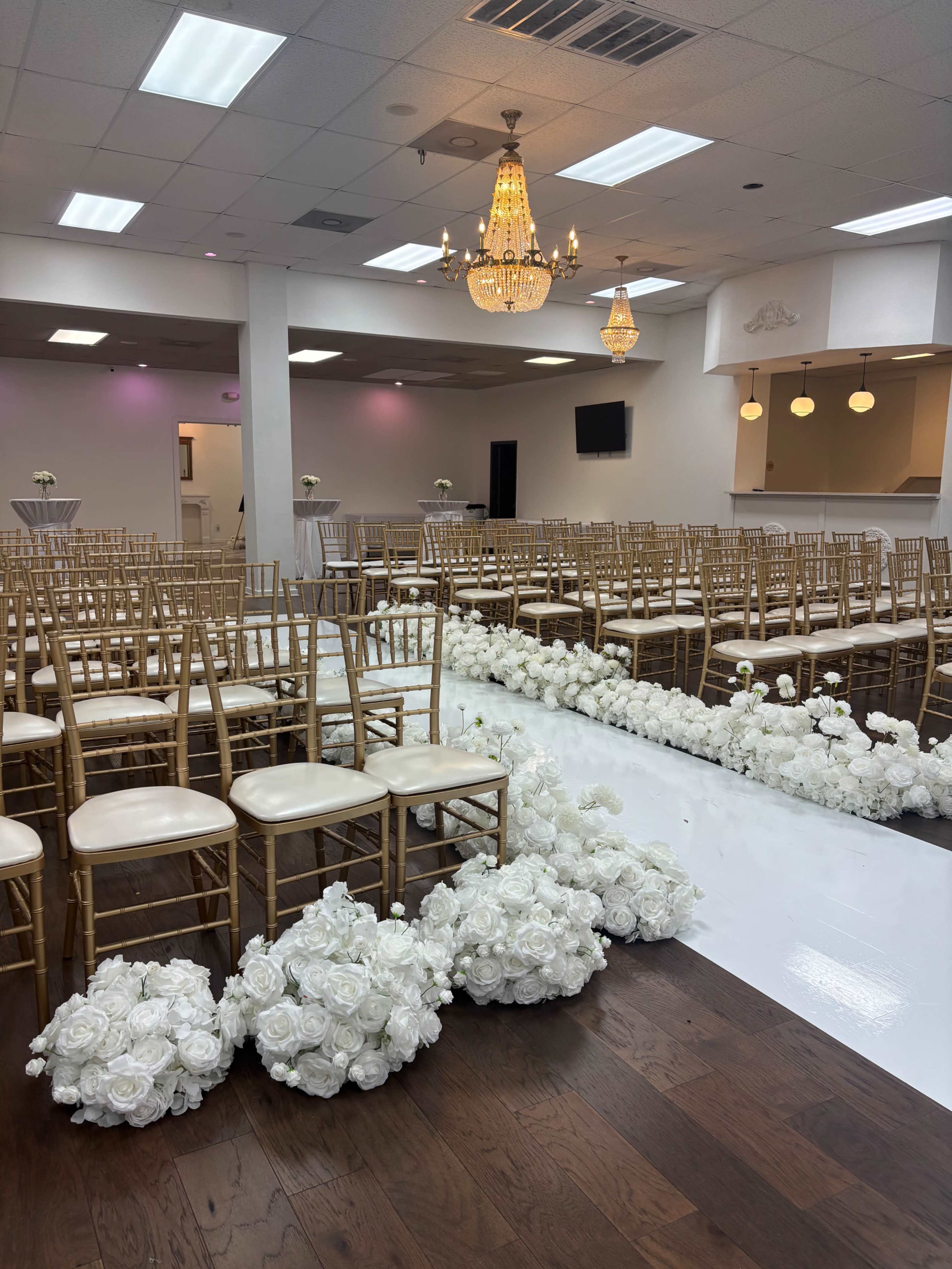 The image depicts a wedding ceremony setup with rows of gold chiavari chairs and white floral arrangements along a polished floor beneath a chandelier in a well-lit space.