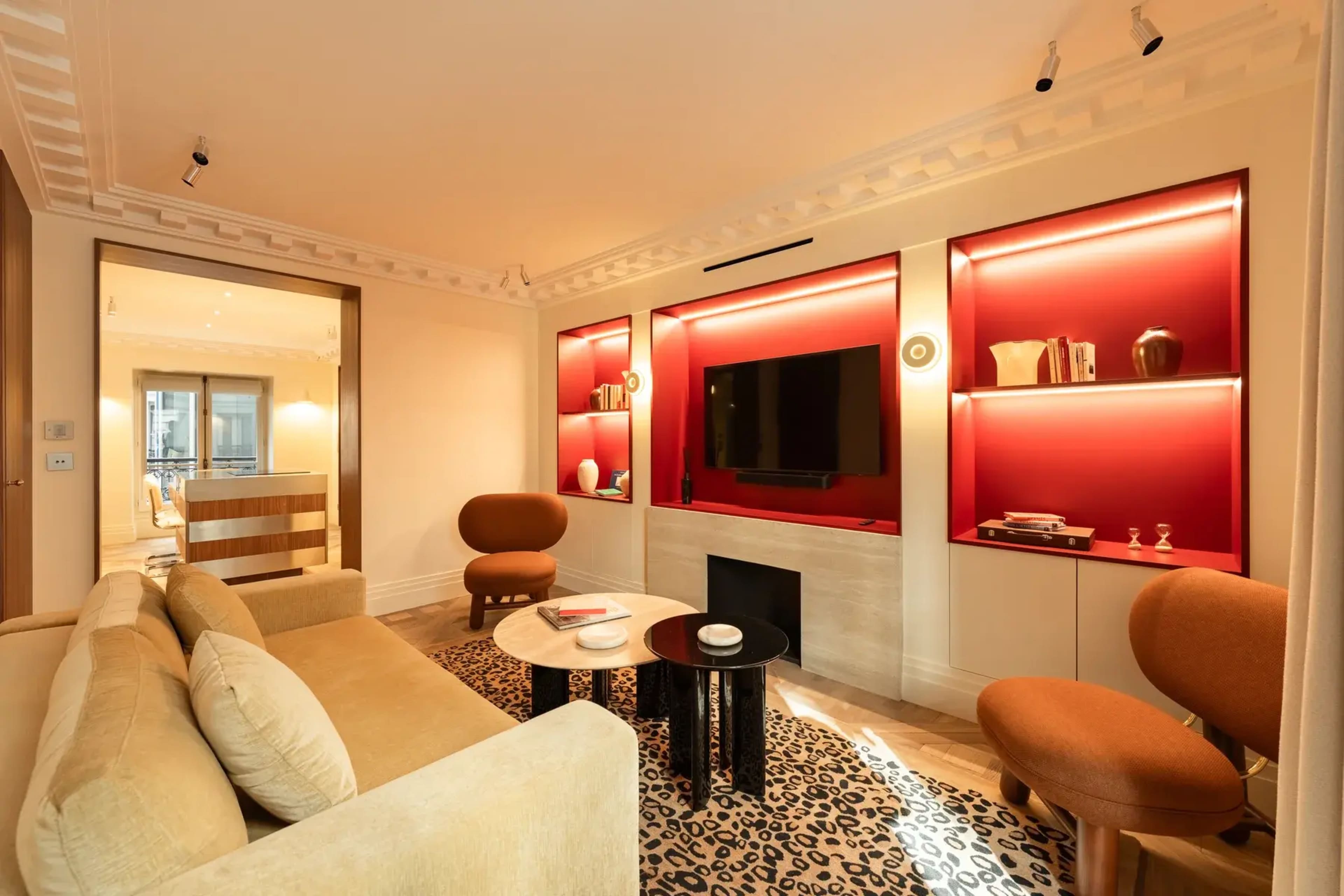 The image shows a modern living room featuring a beige sofa, two orange accent chairs, a coffee table, and a television mounted on a wall with backlit red shelves.
