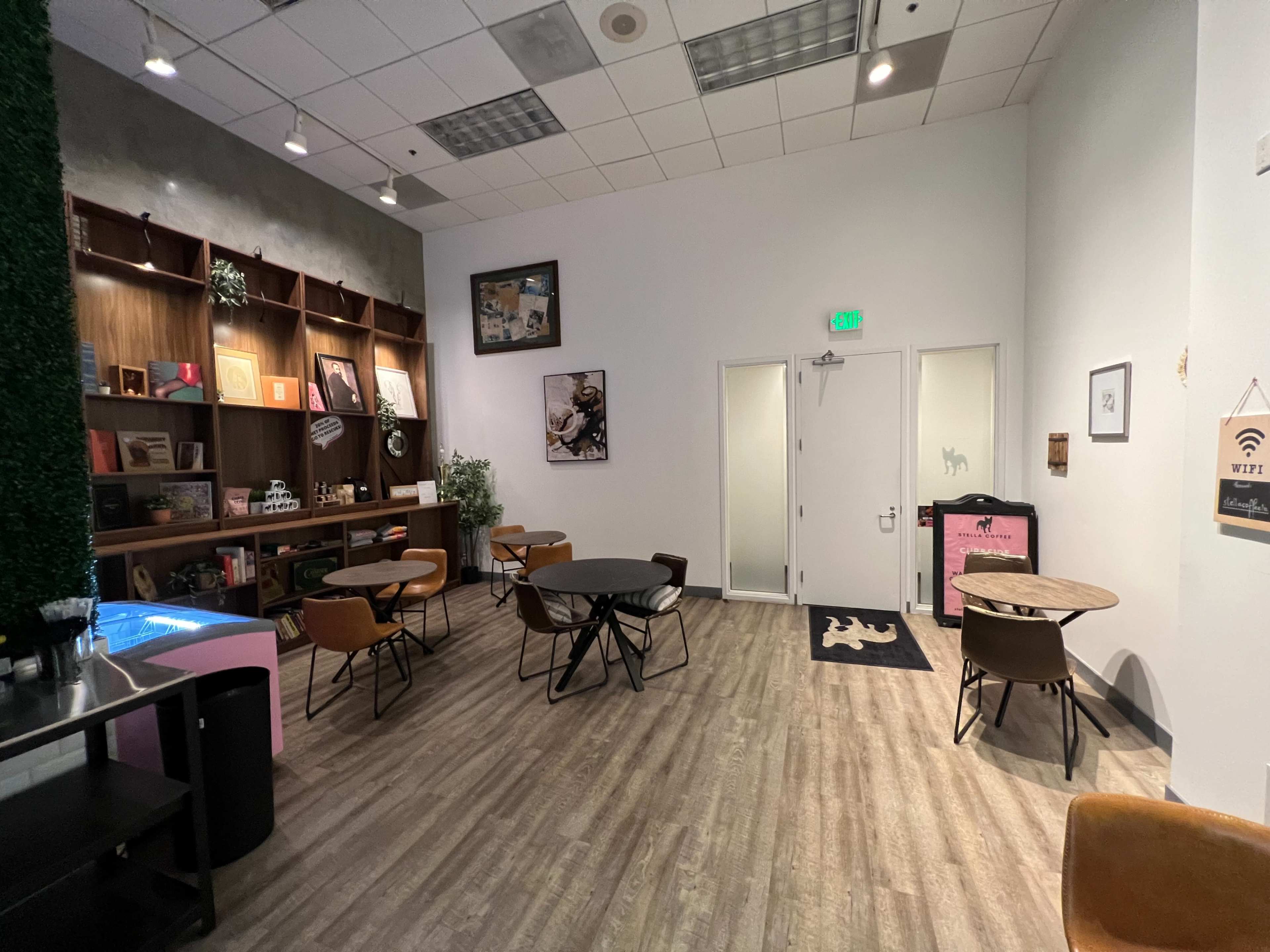 The image shows an indoor café space featuring a seating area with round tables and chairs, a small bar, and decorated walls.