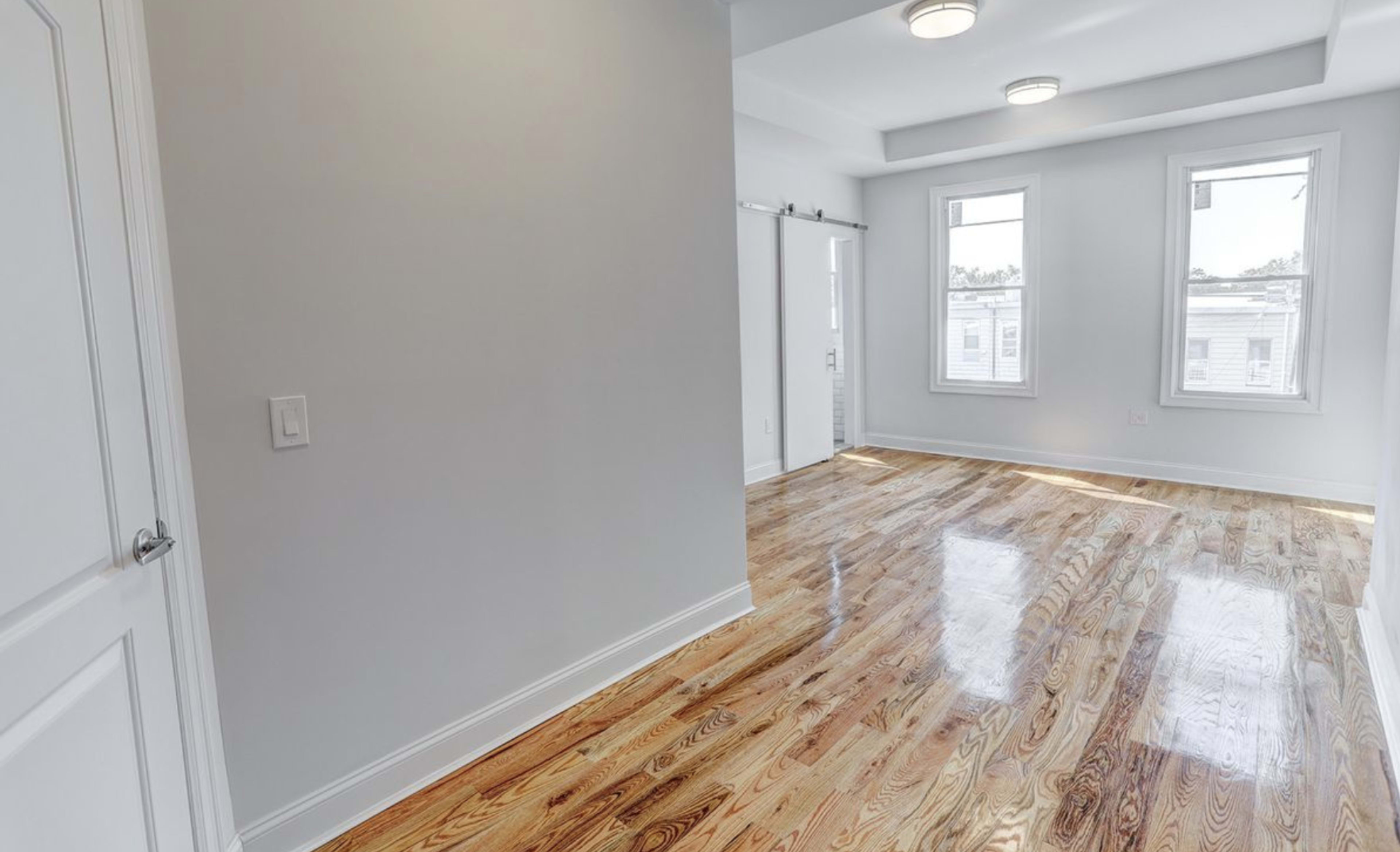 A vacant room with polished wooden floors, white walls, and two windows allowing natural light.