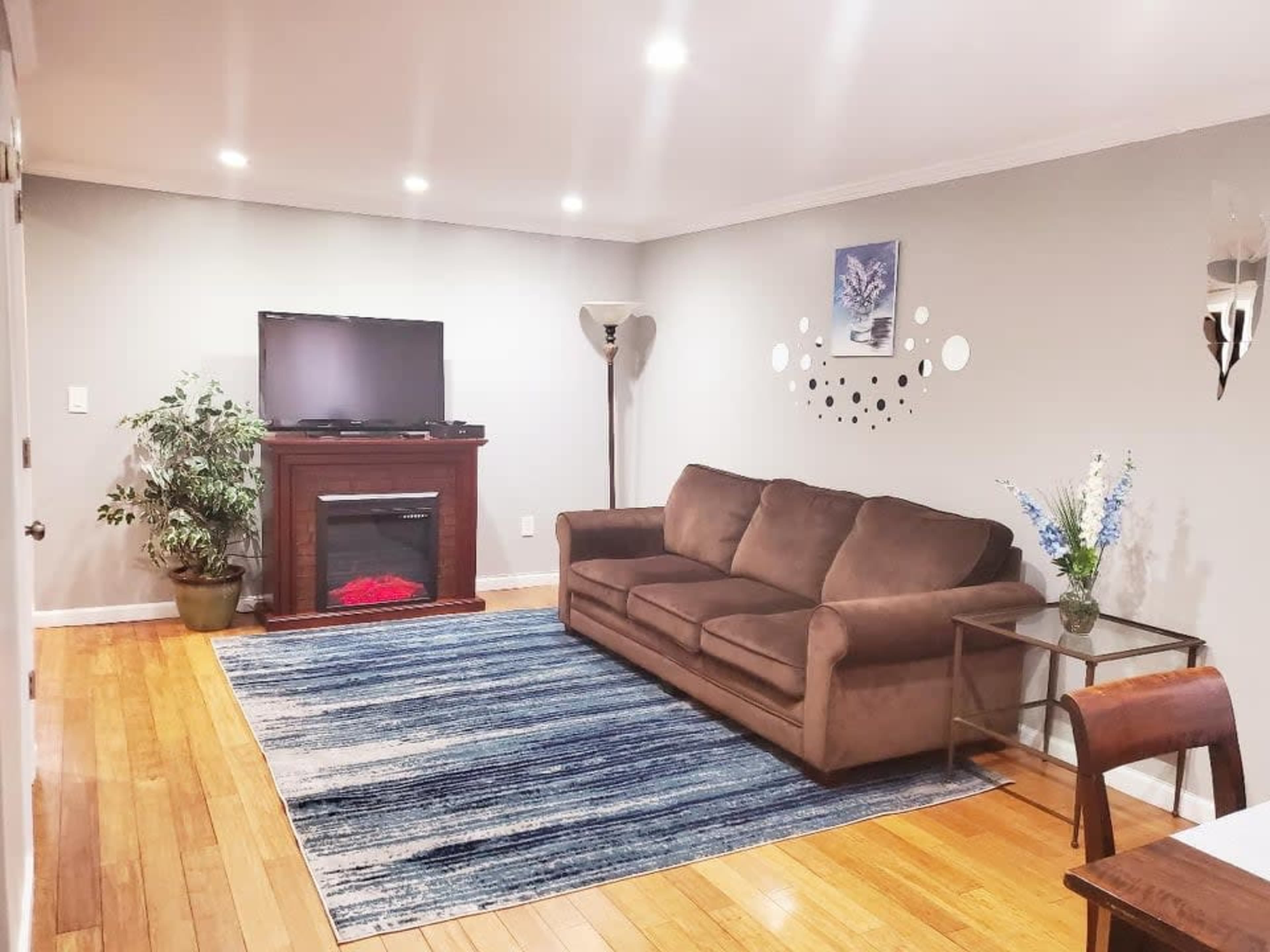 A living room featuring a brown sofa, a television on a wooden stand with an electric fireplace below, a patterned area rug, a small table, and a vase with flowers.