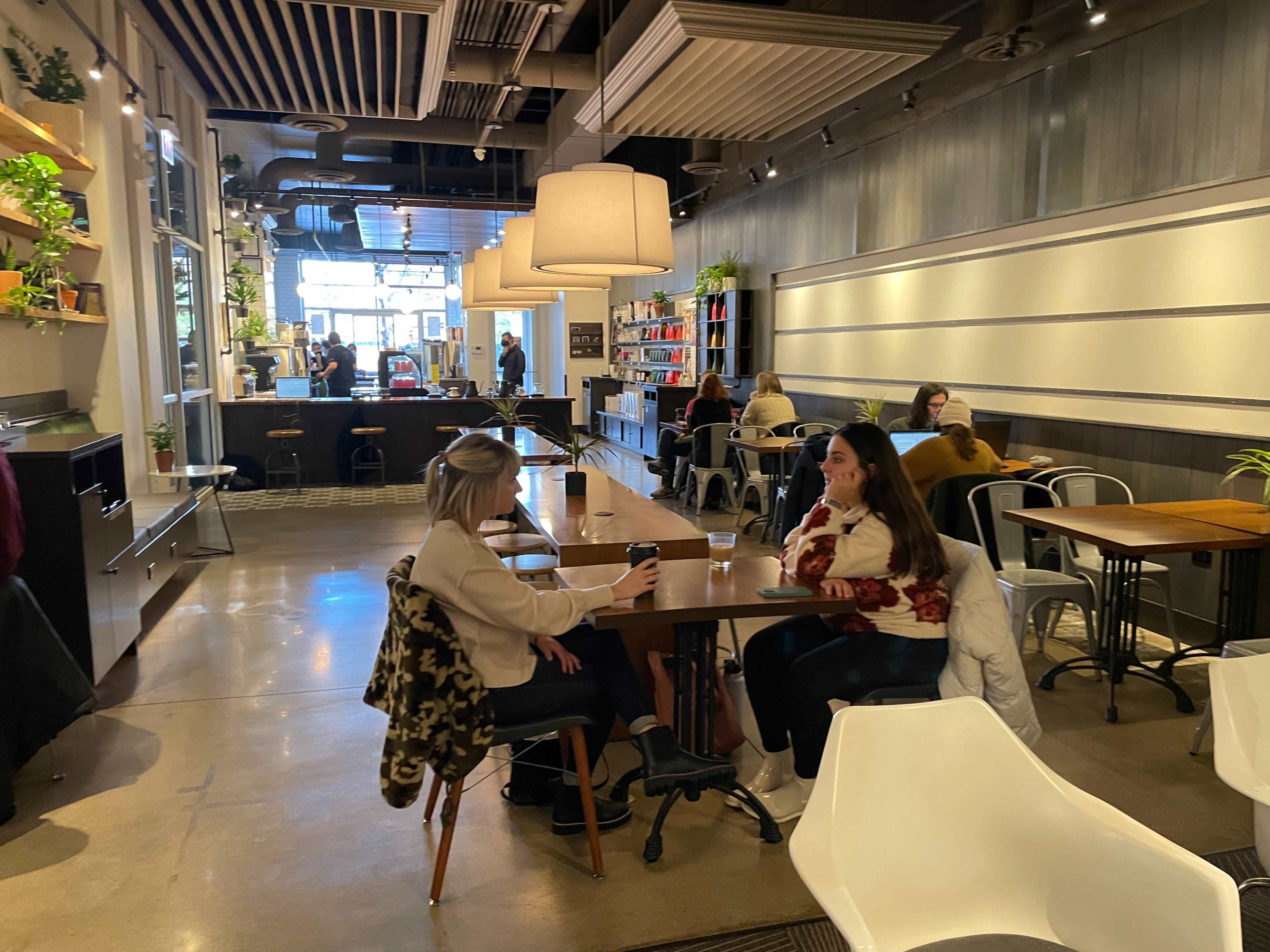 Two women sit at a table in a cafe, while other customers relax at various tables and a barista works behind the counter.