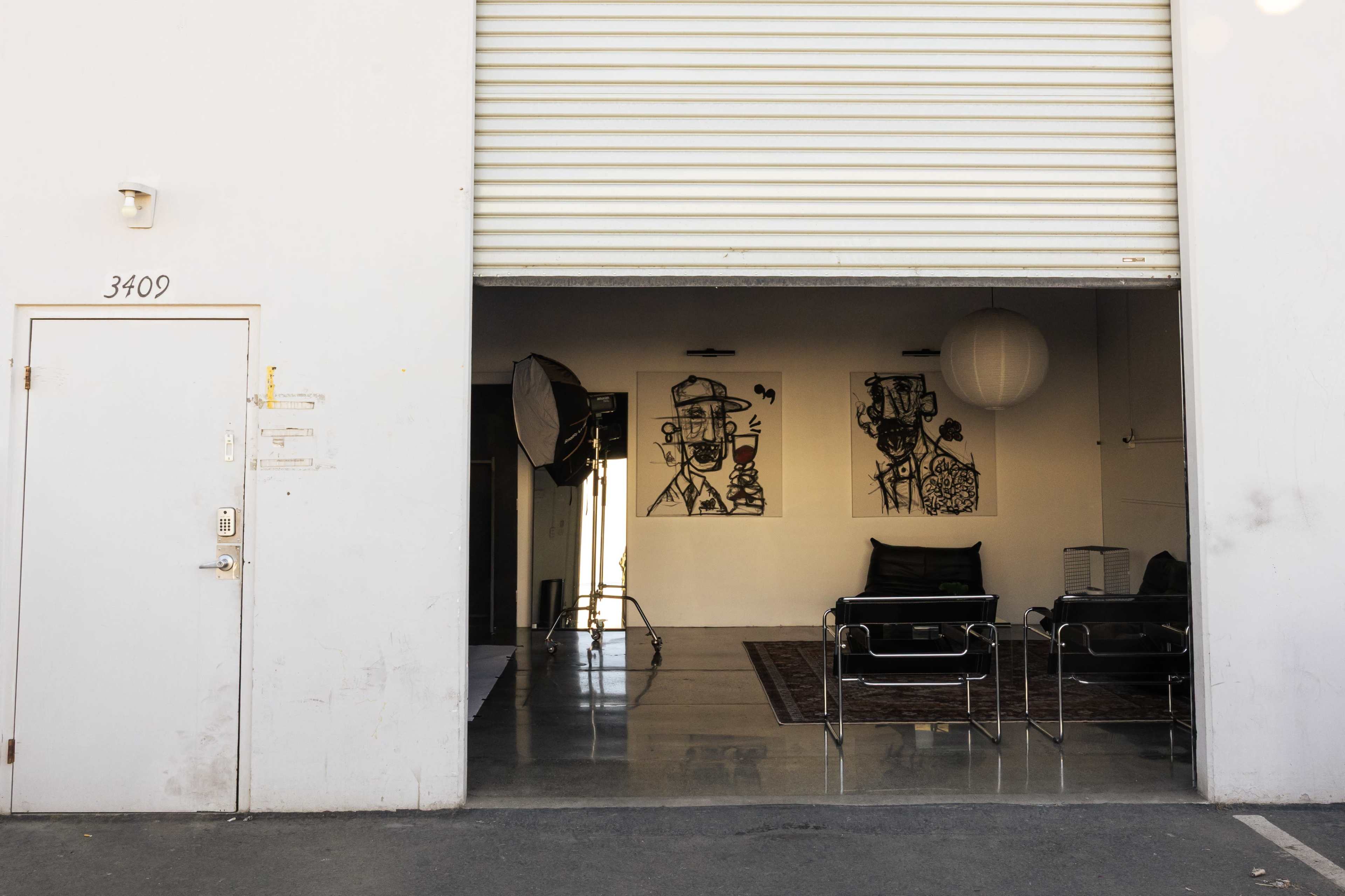 The image shows a glimpse into a studio space with two framed artworks on the wall, a sofa, and a rug on a polished concrete floor, visible through a partially opened garage door.