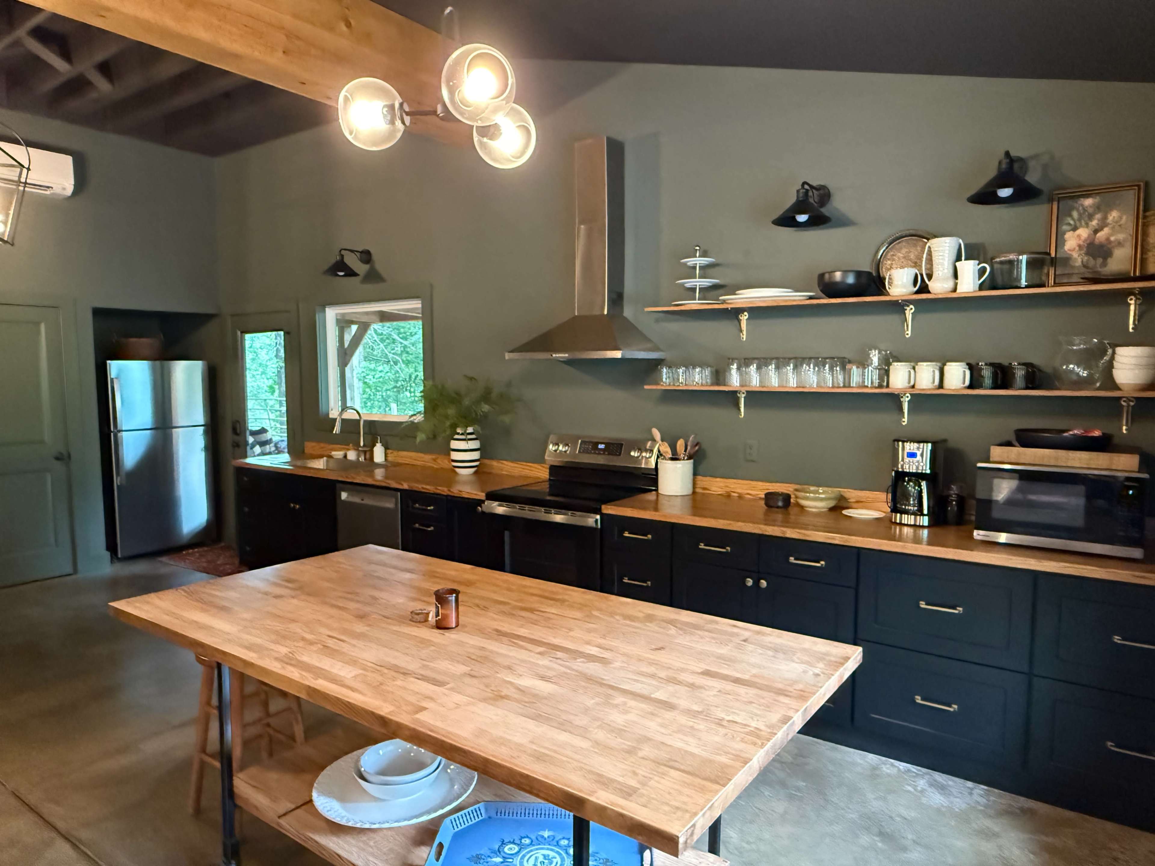 The image shows a modern kitchen with wooden countertops, black cabinetry, and a large central wooden table, featuring stainless steel appliances and various kitchenware displayed on shelves.