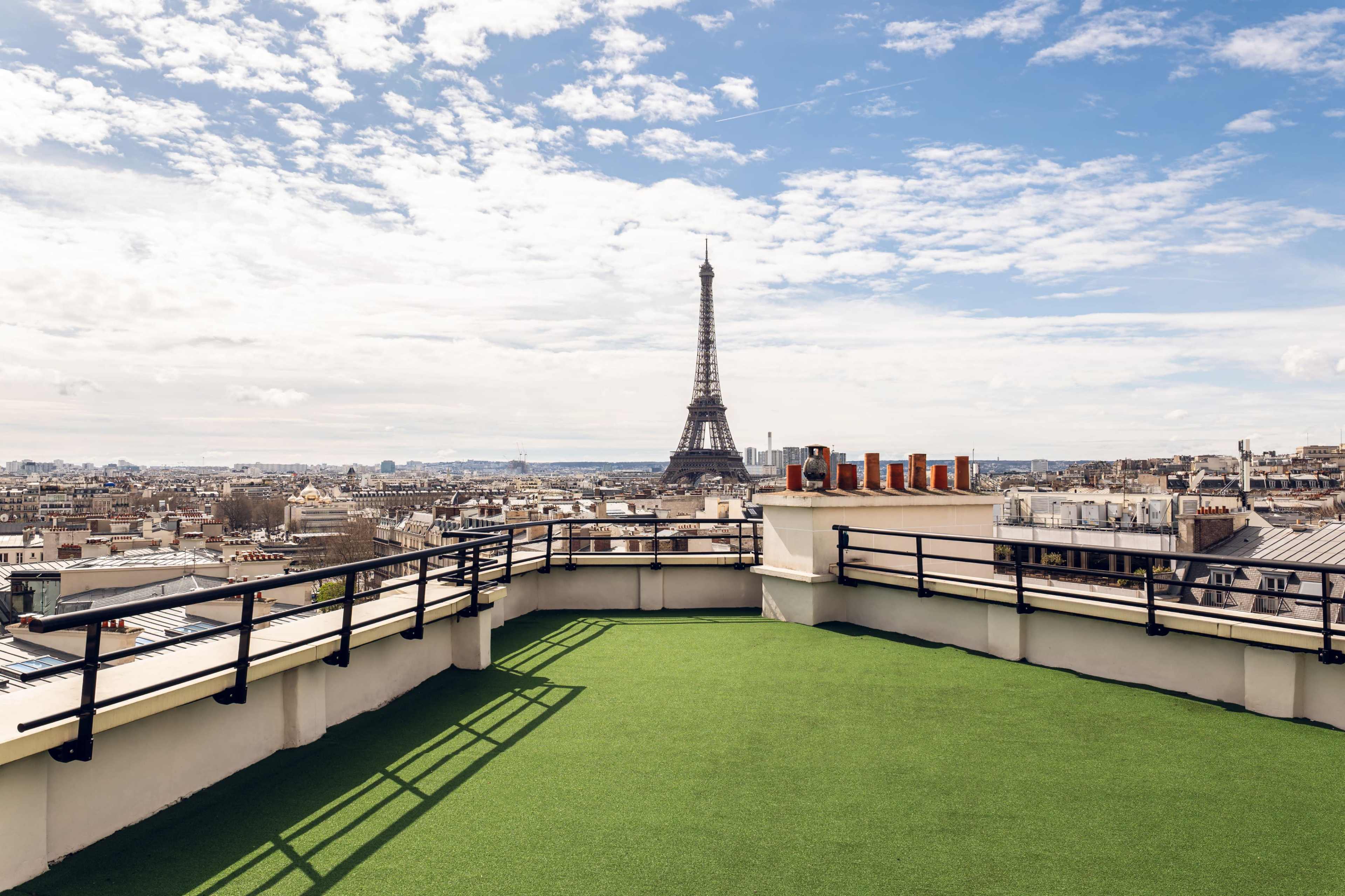 A rooftop terrace with green grass overlooks the Eiffel Tower and the city of Paris under a partly cloudy sky.