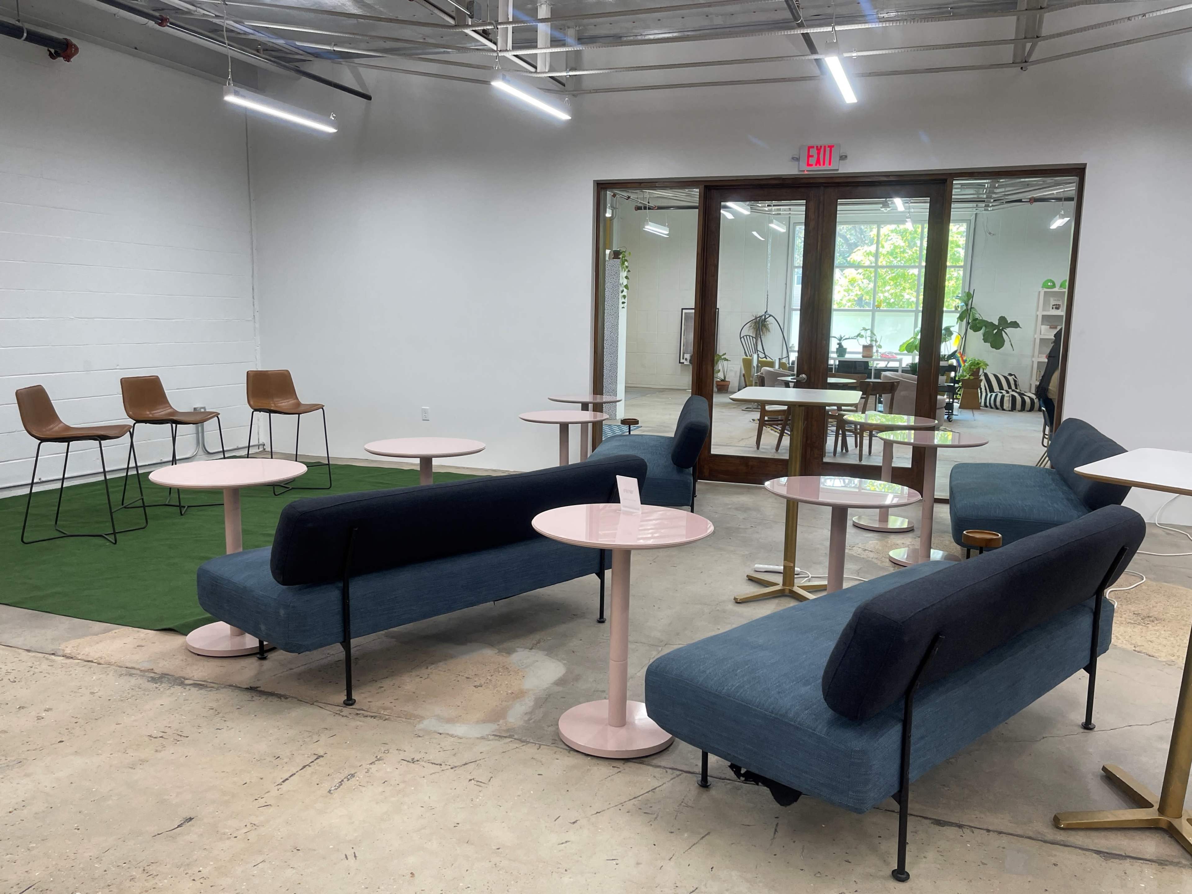 A modern, open lounge area with blue seating, pink tables, and a few wooden chairs, illuminated by natural light coming through large windows.